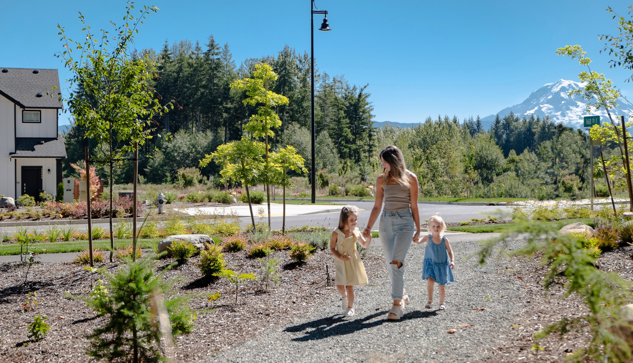 A woman and two young girls are walking on a gravel path surrounded by lush greenery, with a snow-capped mountain visible in the background.