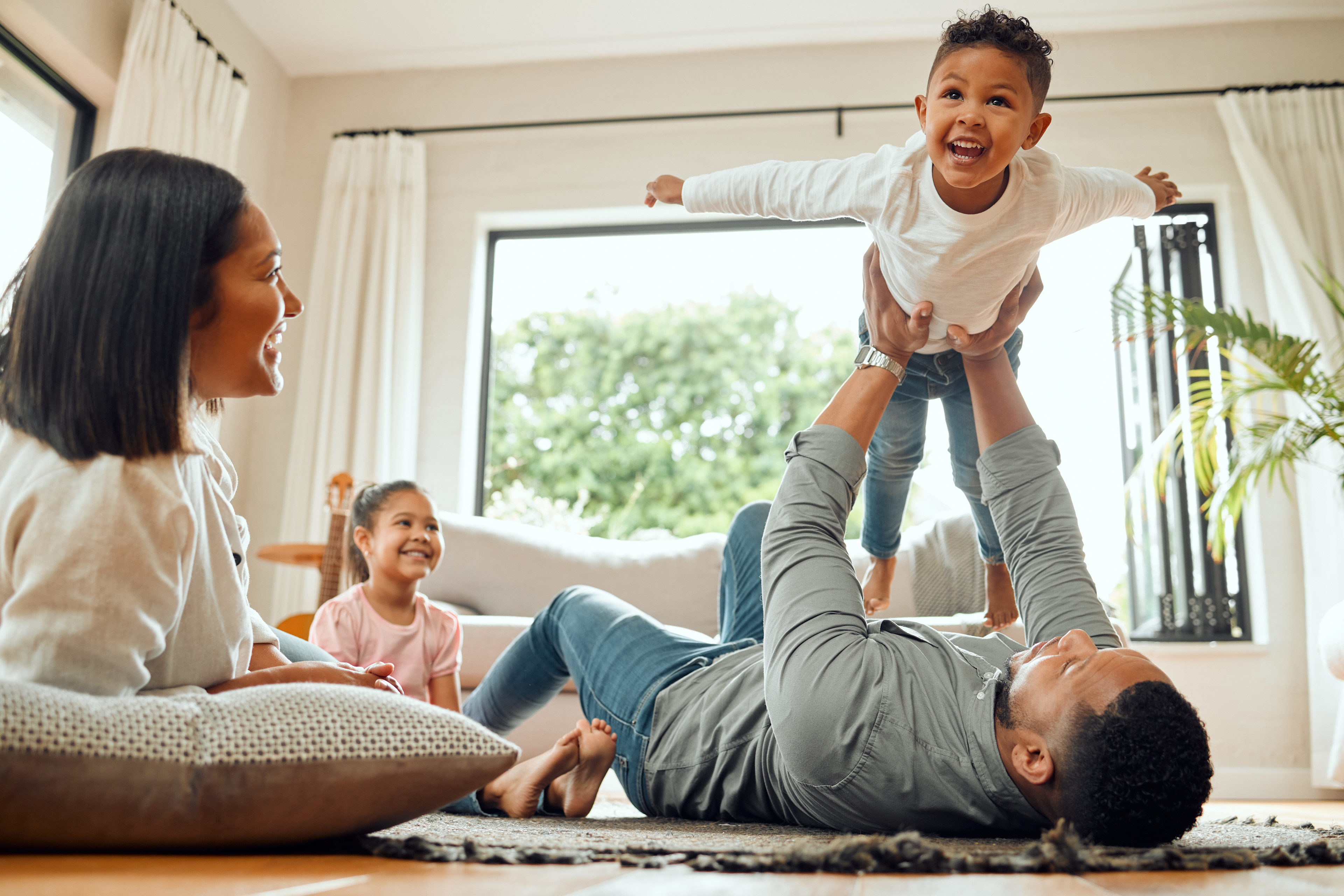 A family enjoying quality time together, with a young child playfully jumping on a parent lying on the floor, while another child sits nearby on a couch.
