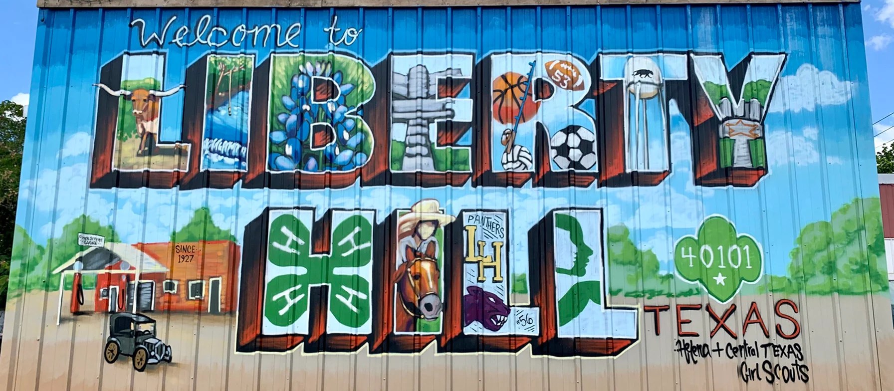 The image depicts a colorful mural with the text "Welcome to Liberty, Texas" prominently displayed. The mural features various symbols and icons representing the town, including a soccer ball, a cowboy hat, and a four-leaf clover.