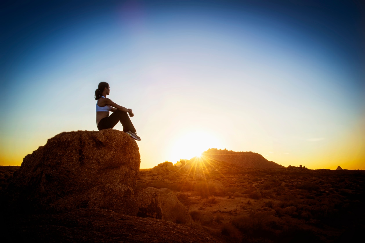 A silhouetted figure sits atop a rocky outcrop, overlooking a stunning desert landscape bathed in the warm glow of the setting sun.
