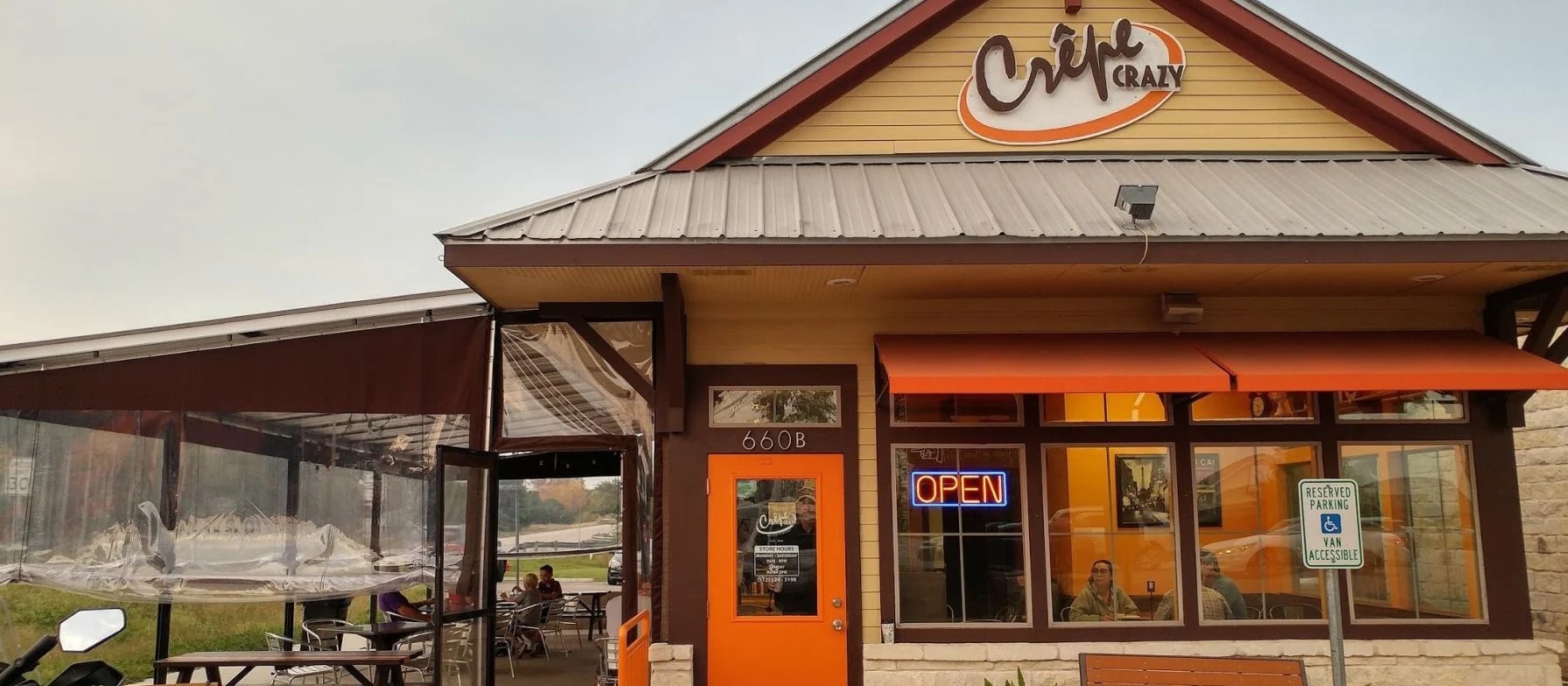 A cozy-looking cafe with a wooden exterior, an open sign, and people sitting at tables in the foreground.