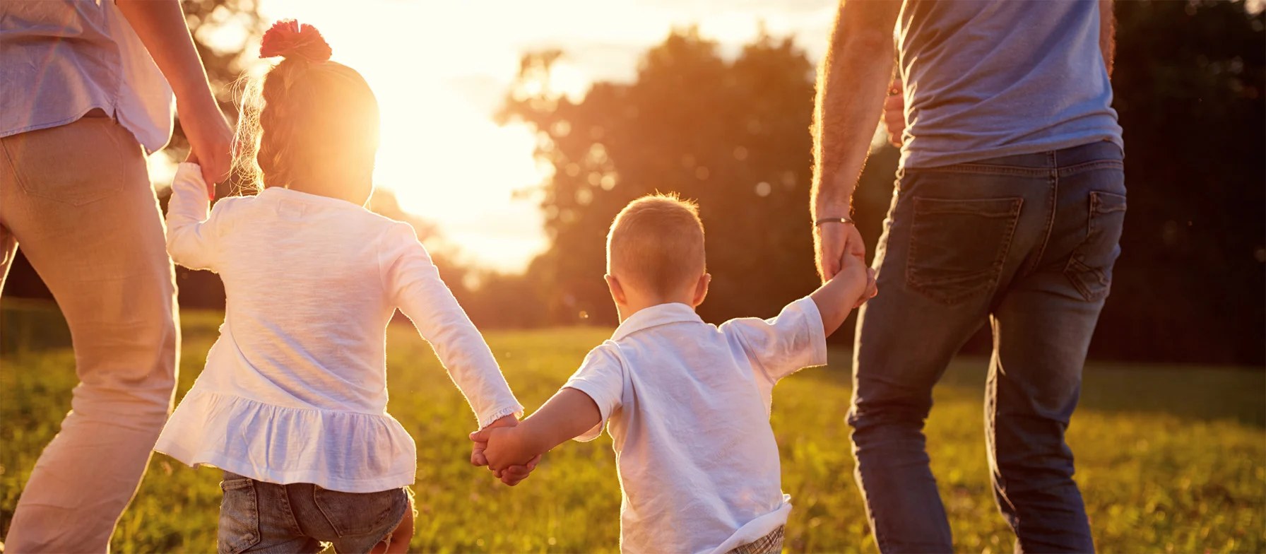 A group of people, likely a family, walking hand-in-hand through a serene, sun-dappled field, with a backdrop of lush greenery and a warm, golden glow.