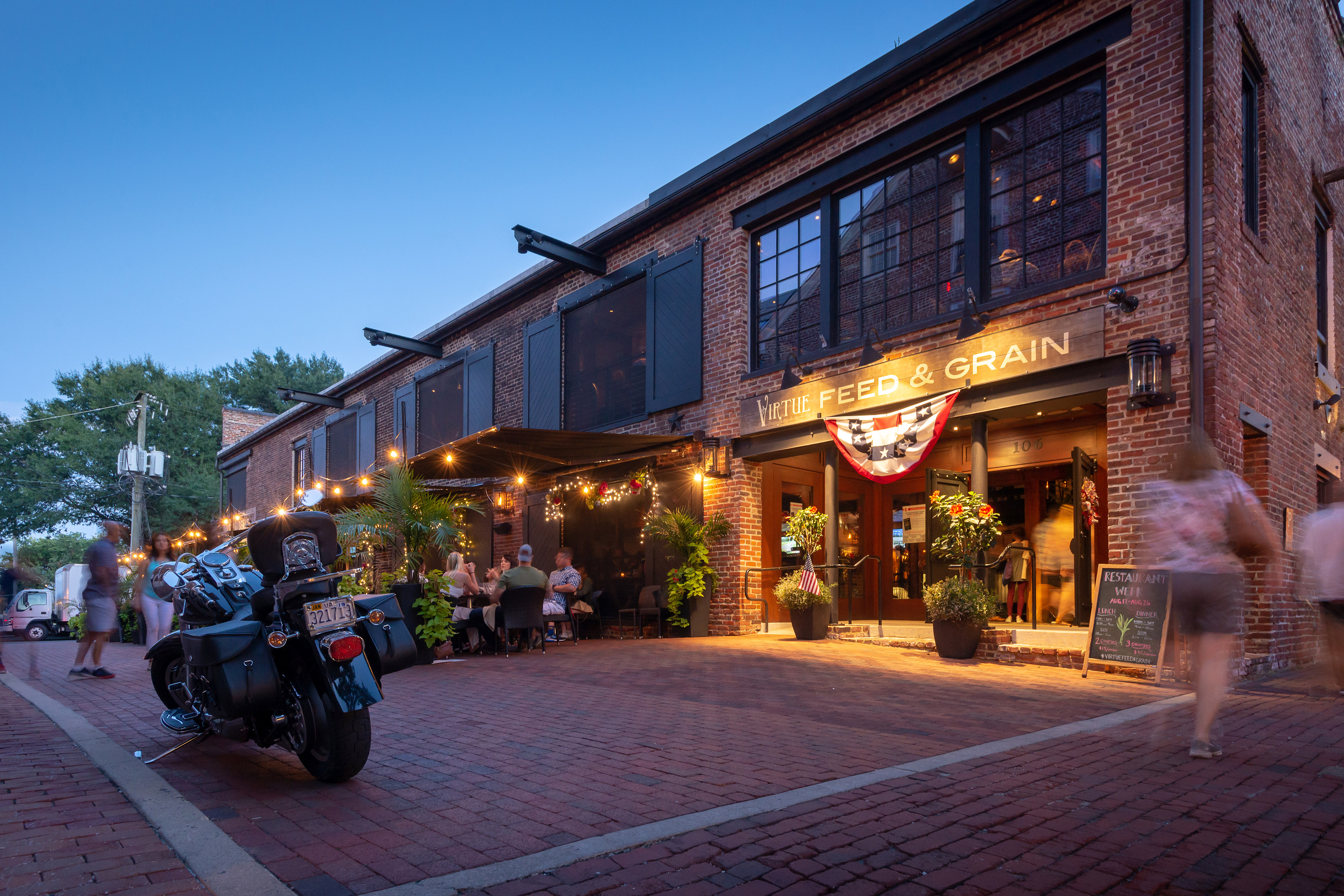 A lively street scene with a brick building housing a restaurant called "Seed & Grain" and several people walking along the sidewalk, with a motorcycle parked in the foreground.