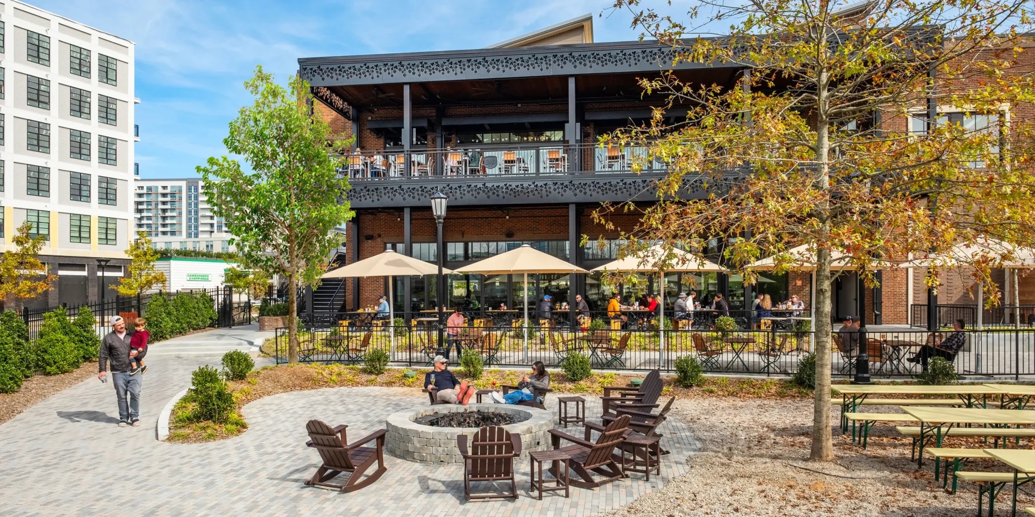 A cozy outdoor seating area with wooden furniture and umbrellas, surrounded by trees and buildings in the background, creating a pleasant and inviting atmosphere.