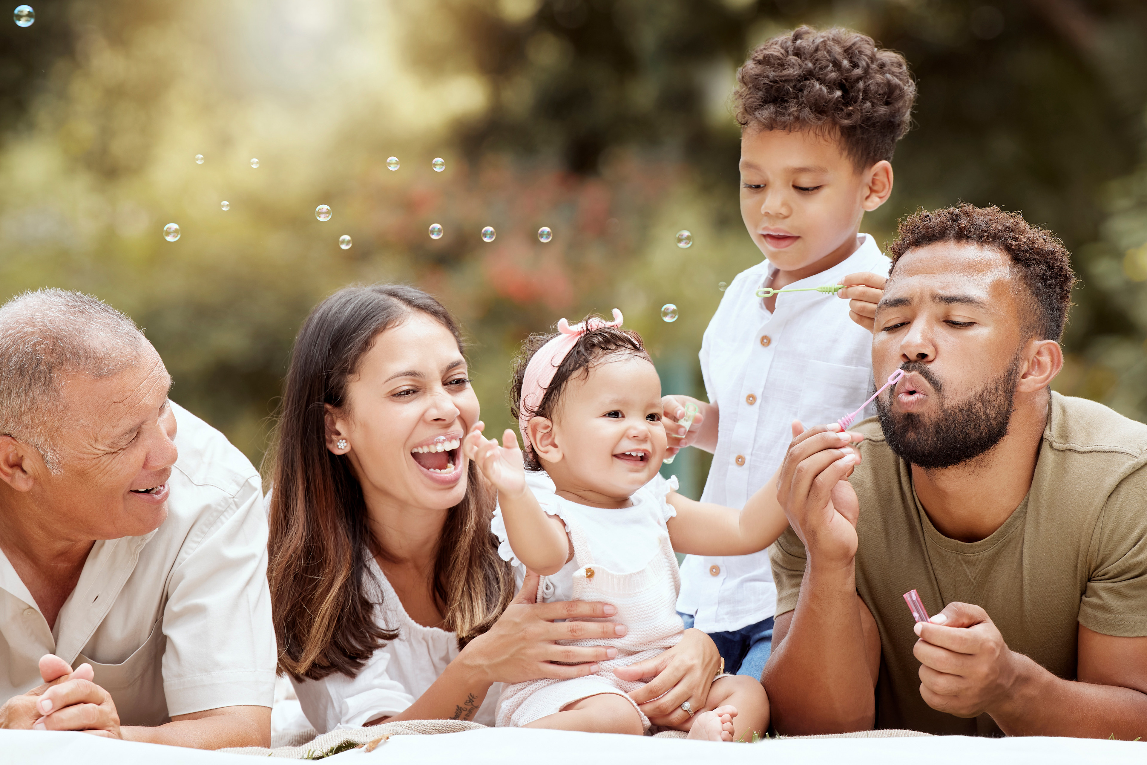 A happy, diverse family sitting together outdoors, surrounded by a blurred natural setting with sunlight filtering through.