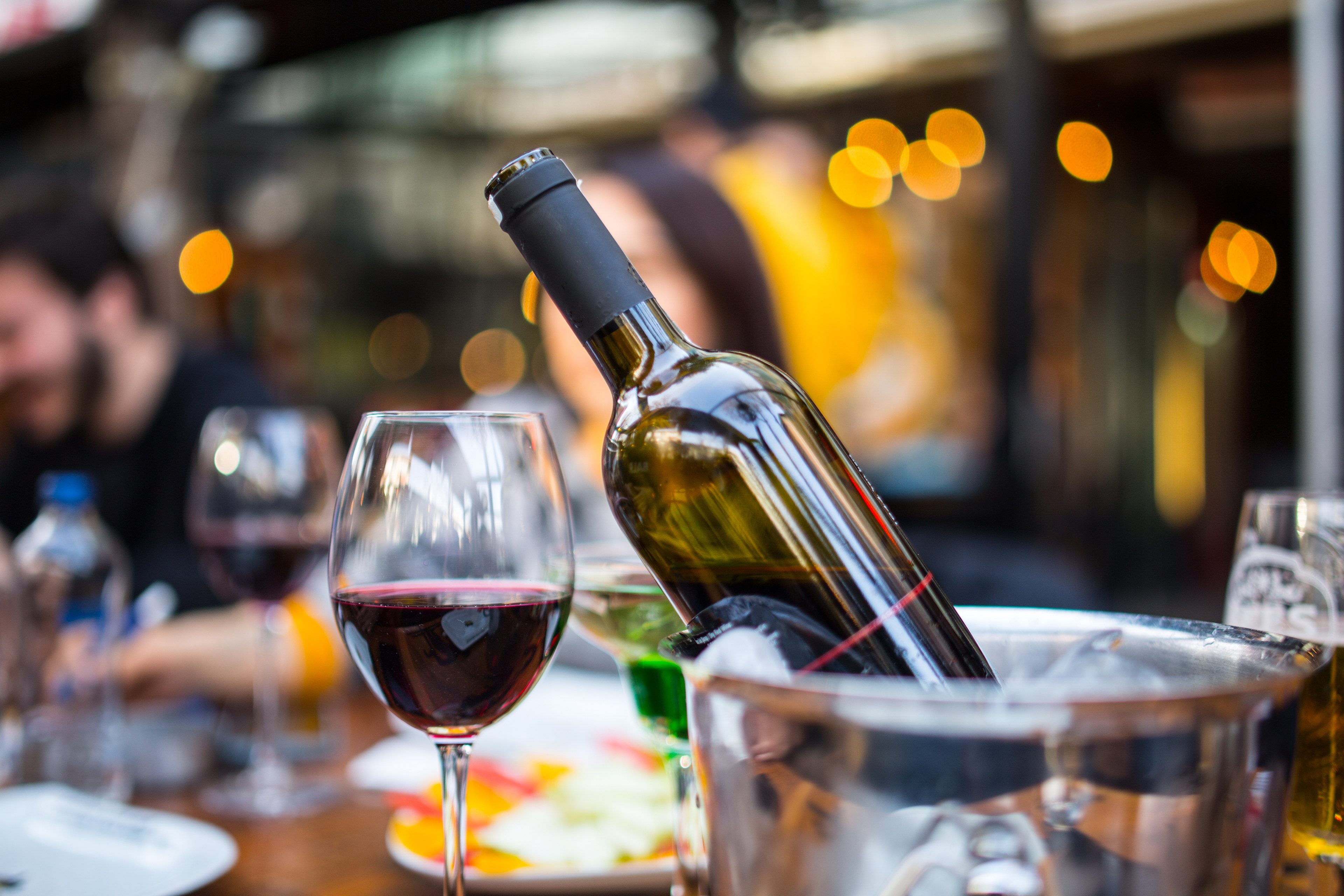 A bottle of wine and a glass of red wine are prominently displayed in the foreground, with a blurred background of what appears to be a restaurant or bar setting with colorful lights.
