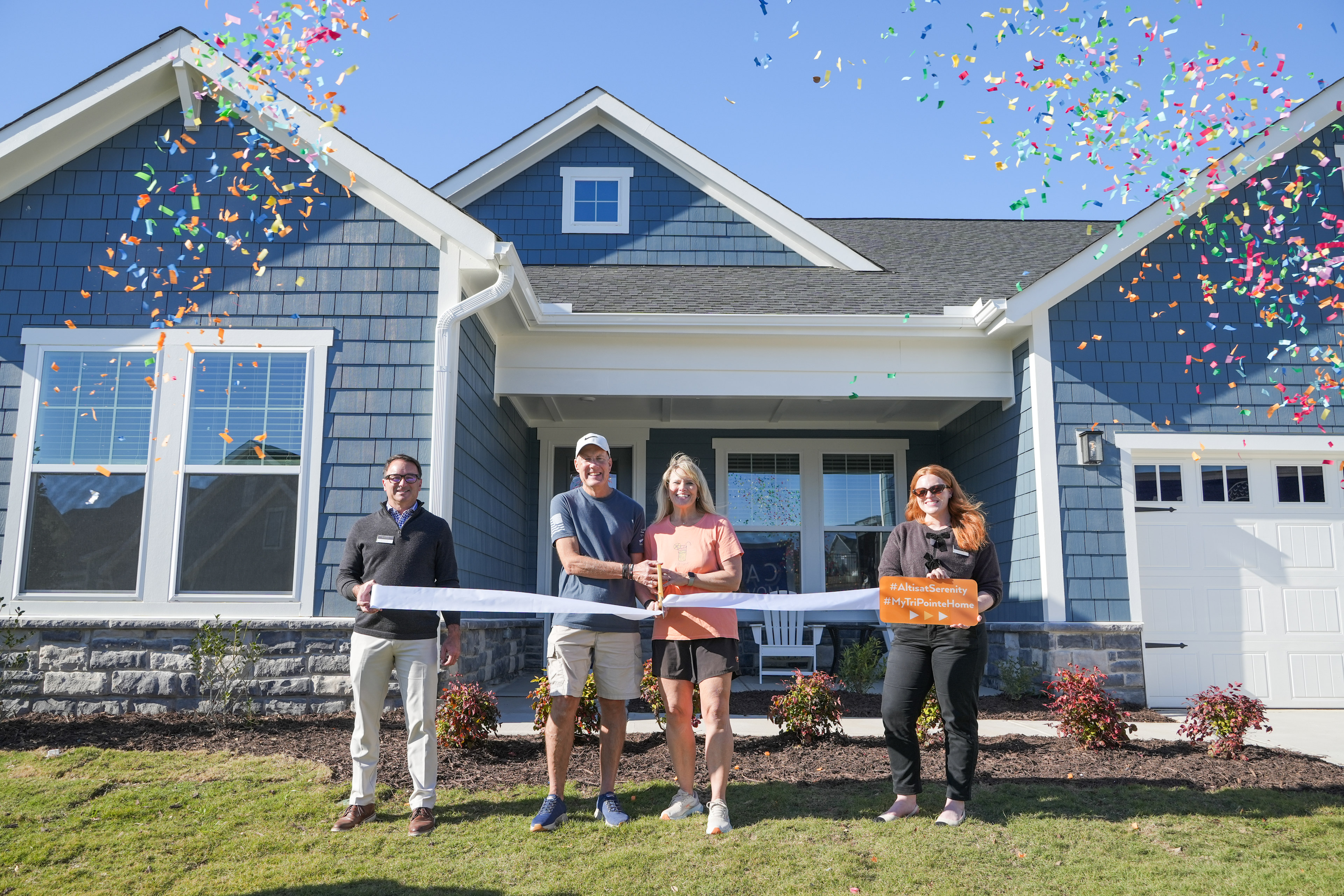 A group of four people standing in front of a newly constructed blue and white house, surrounded by colorful confetti in the air.