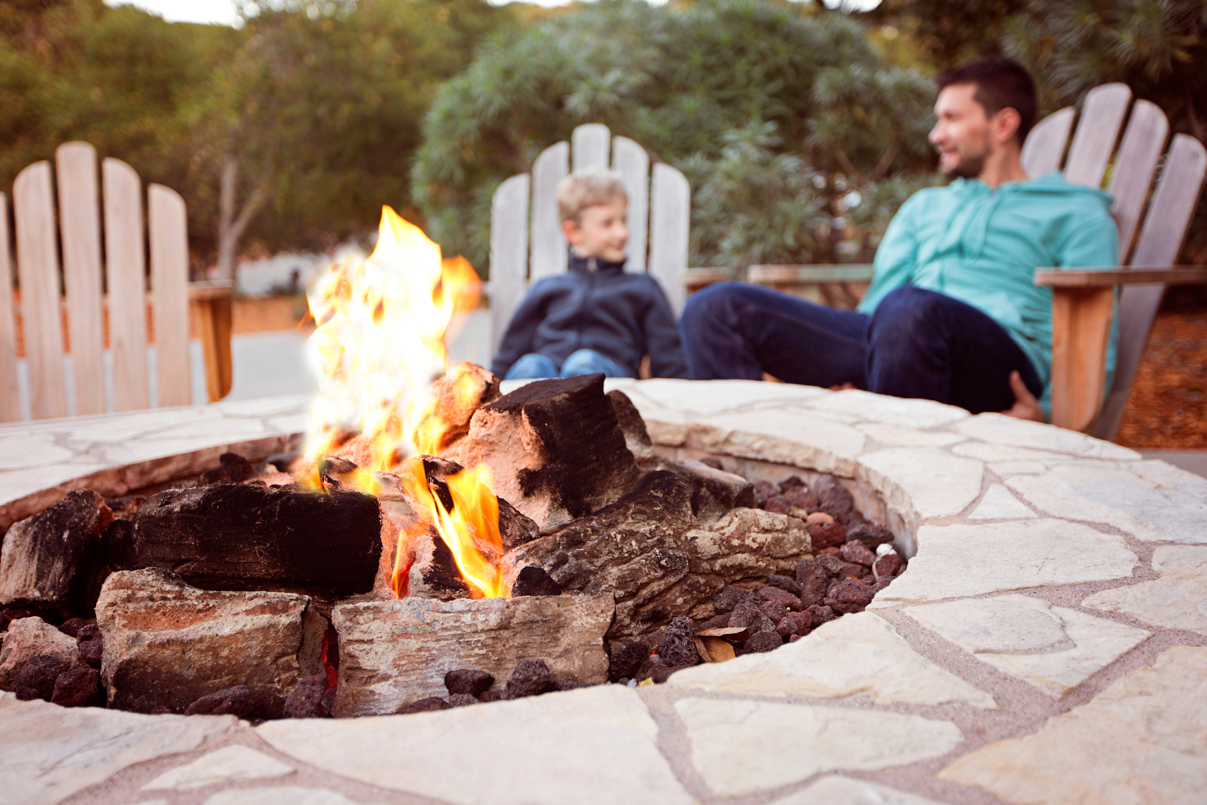 A cozy outdoor fire pit surrounded by stone pavers, with two people sitting on wooden chairs in the background enjoying the warmth and ambiance.
