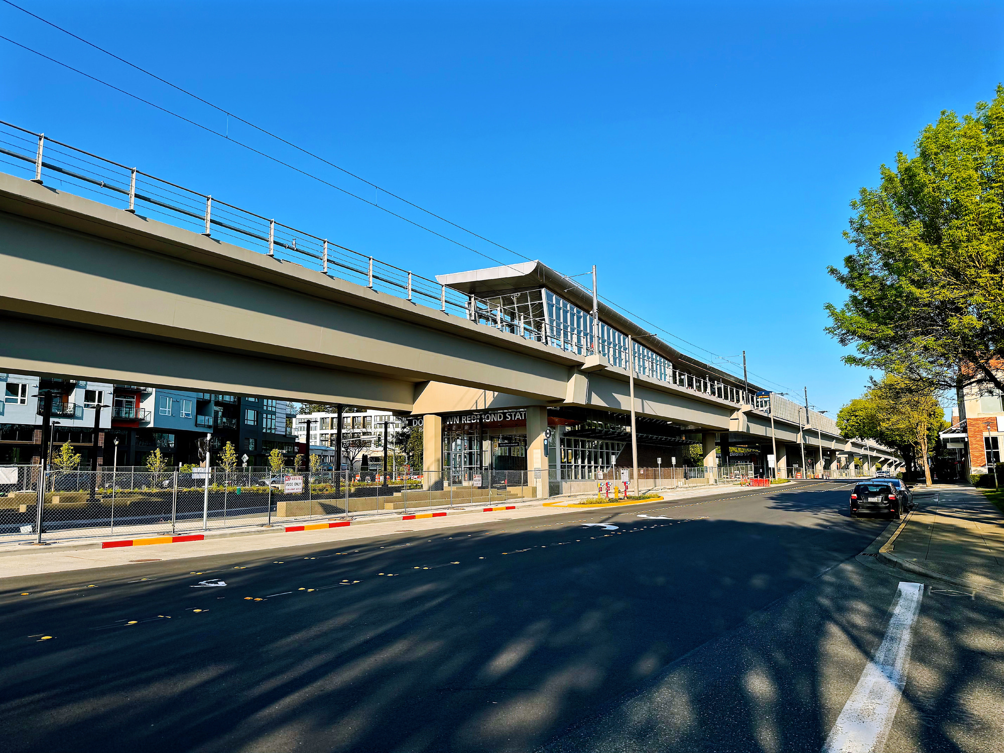 A multi-story building with a raised walkway and shops on the ground level, set against a clear blue sky with trees lining the street in the foreground.