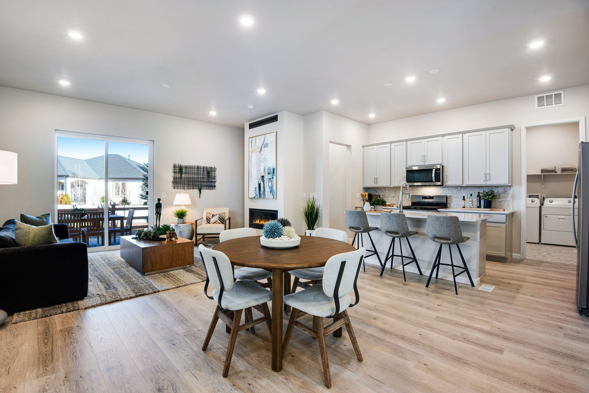 A modern and open-concept living space with a dining area, kitchen, and living room, featuring a wooden dining table, white chairs, and a large window overlooking a scenic outdoor view.