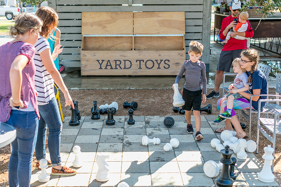 A group of children playing with various outdoor toys and games on a paved area, with a large "Yard Toys" sign in the background.