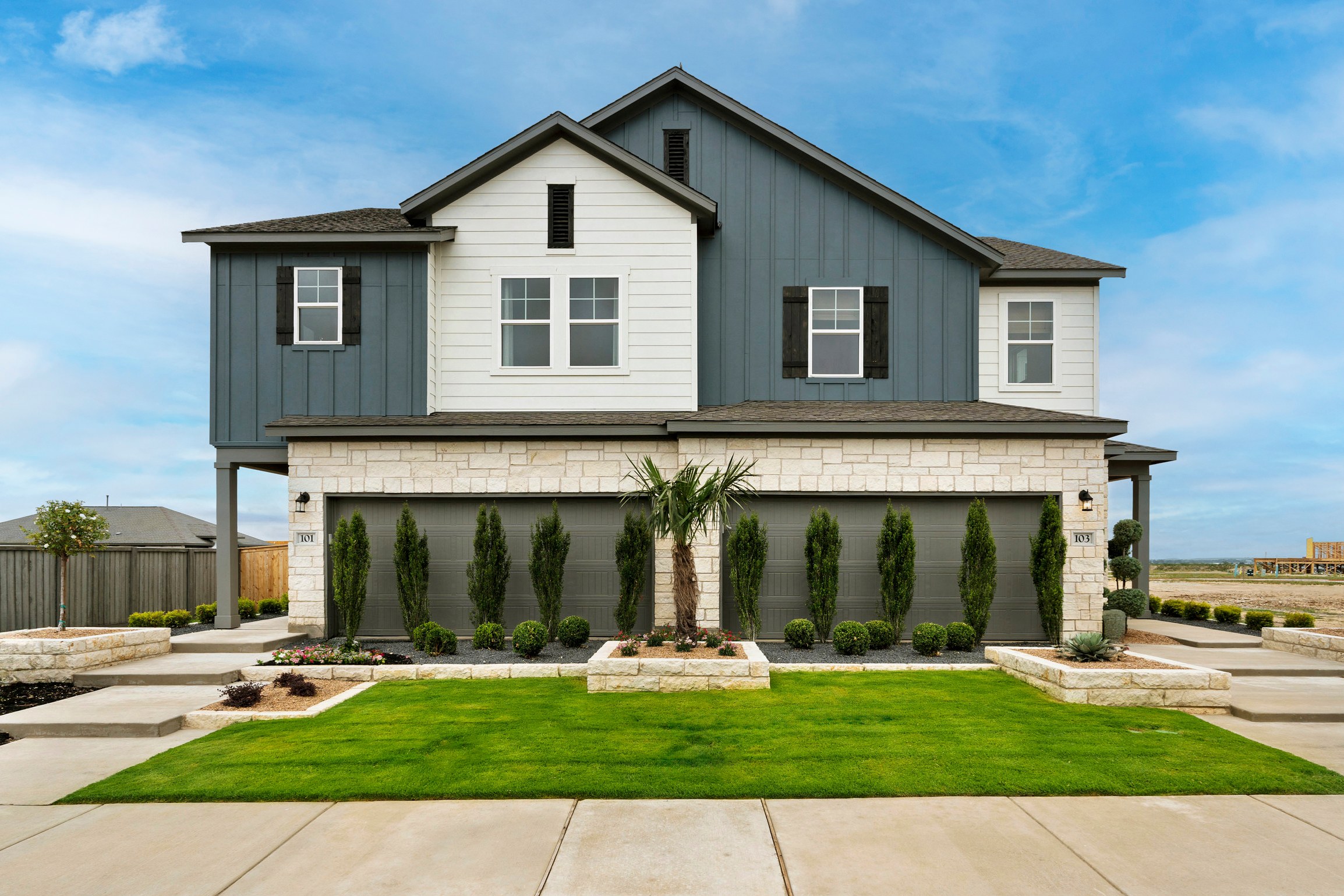 A two-story house with a gray exterior, a well-manicured lawn, and a row of tall evergreen trees in the foreground, set against a clear blue sky.