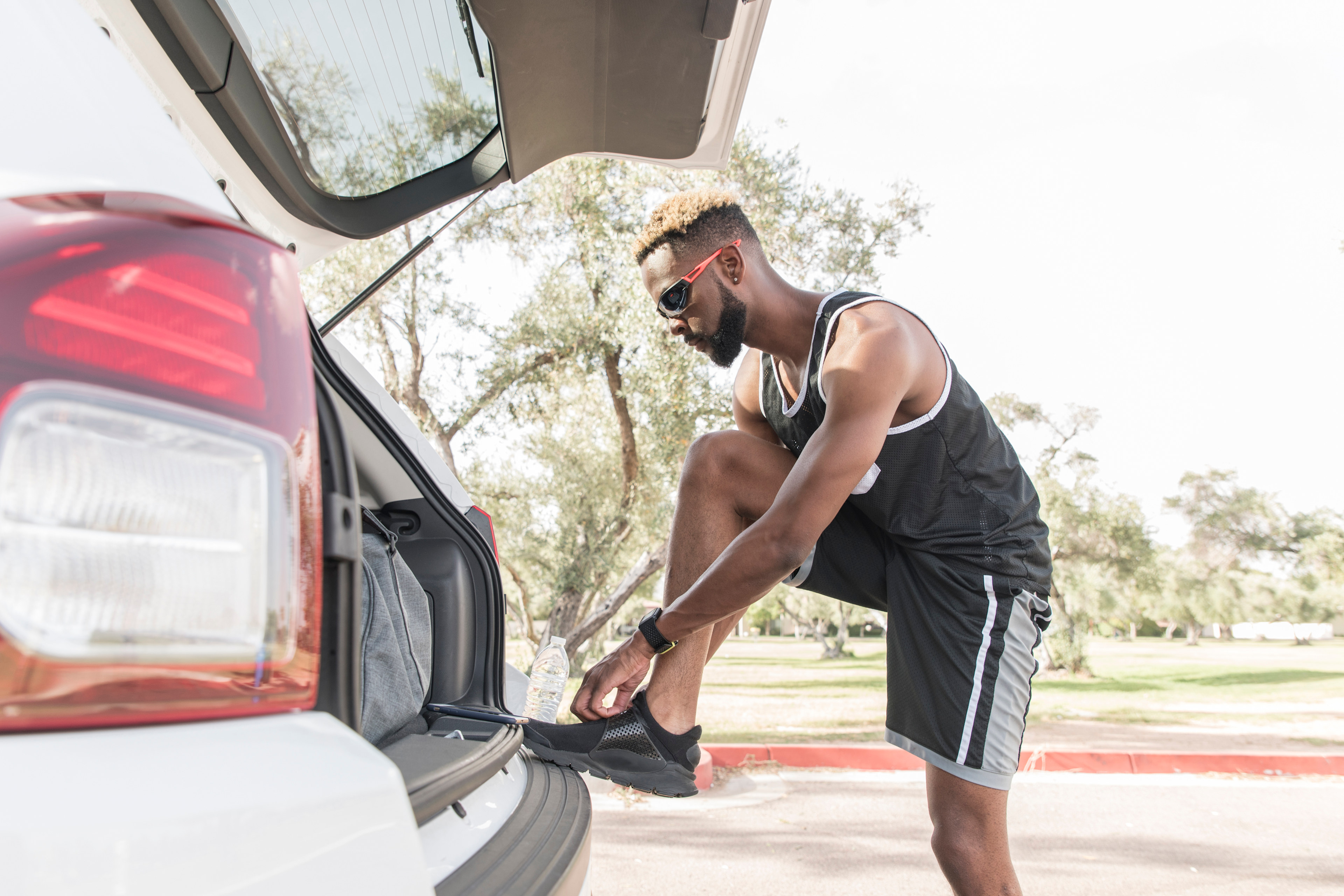 A man in athletic clothing is leaning into the open trunk of a red car, surrounded by a grassy outdoor setting.