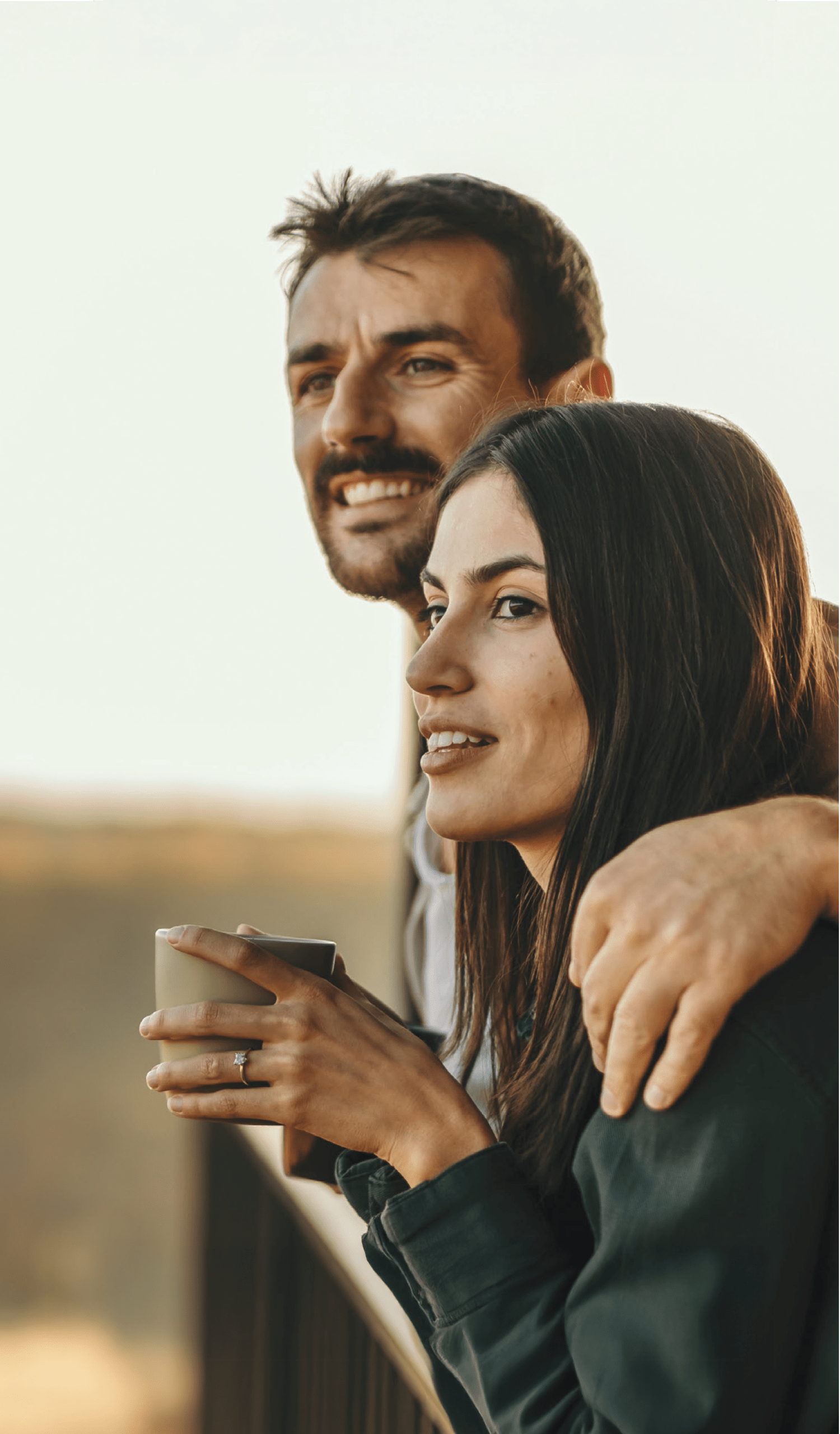 A smiling couple embracing and enjoying the view from a balcony or terrace, with a scenic landscape in the background.