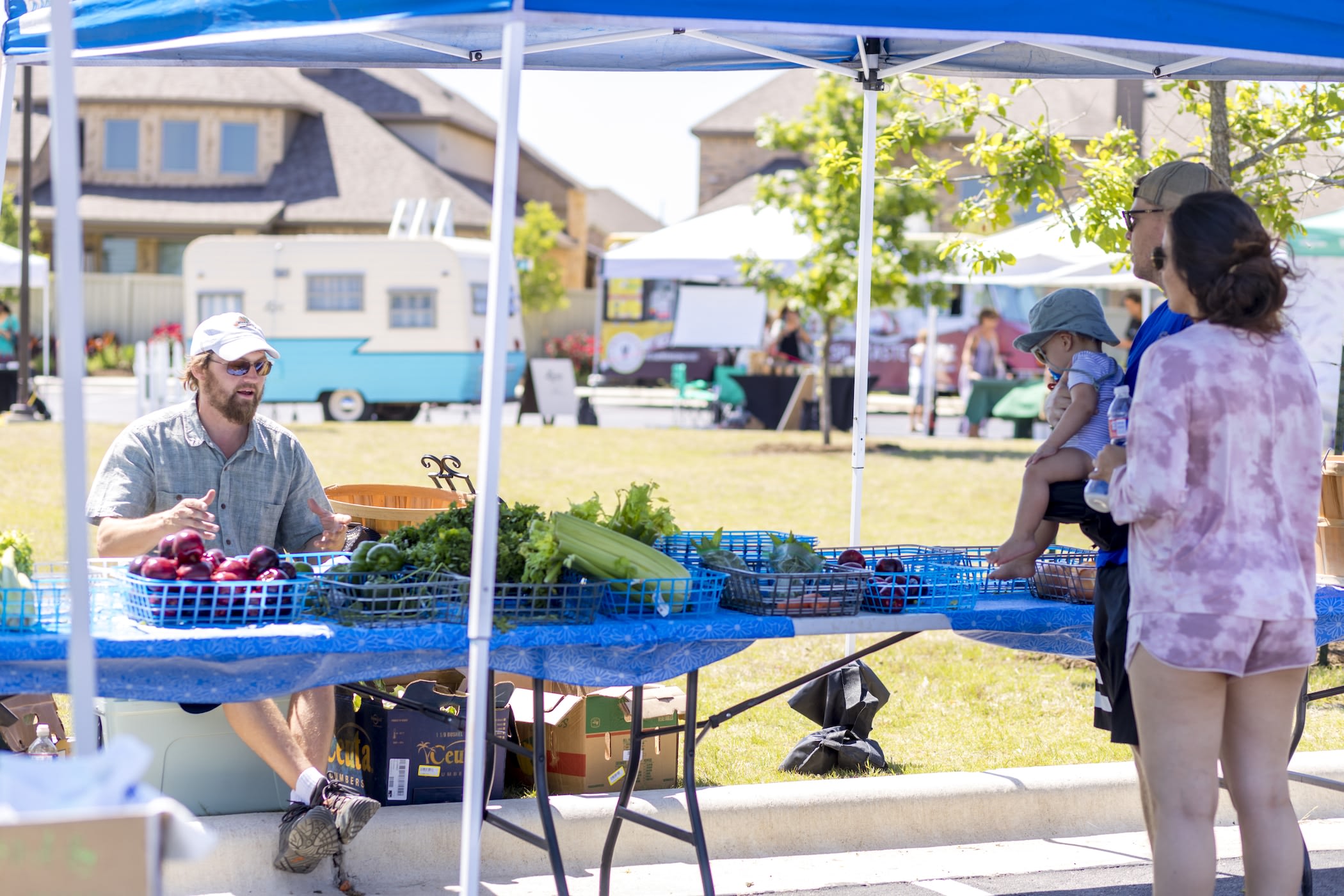 A farmer's market or outdoor market setting with a vendor selling fresh produce from a table, surrounded by people browsing and shopping in the background.