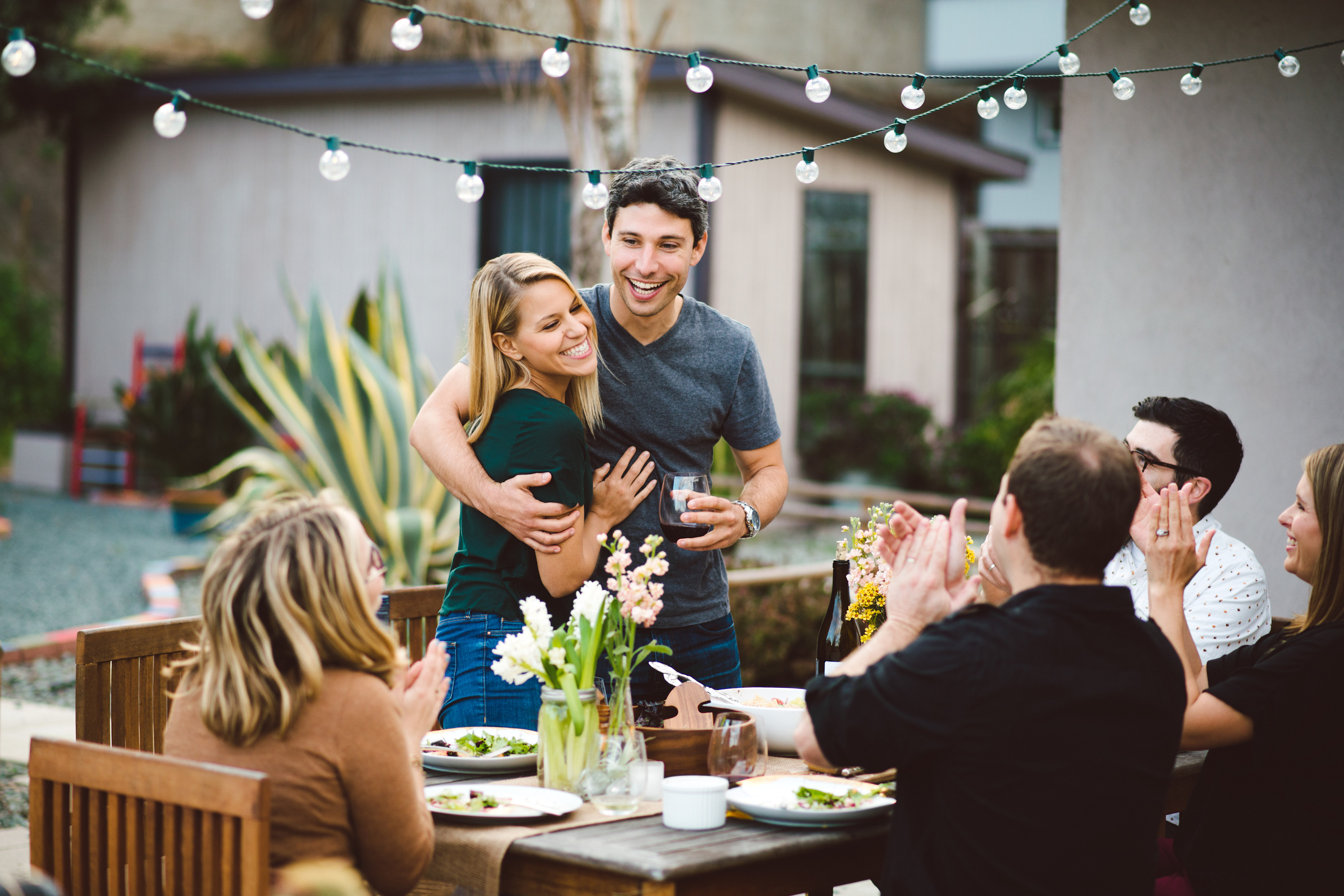 A group of people enjoying an outdoor gathering, with a couple embracing in the foreground and others seated around a table set with food and drinks.