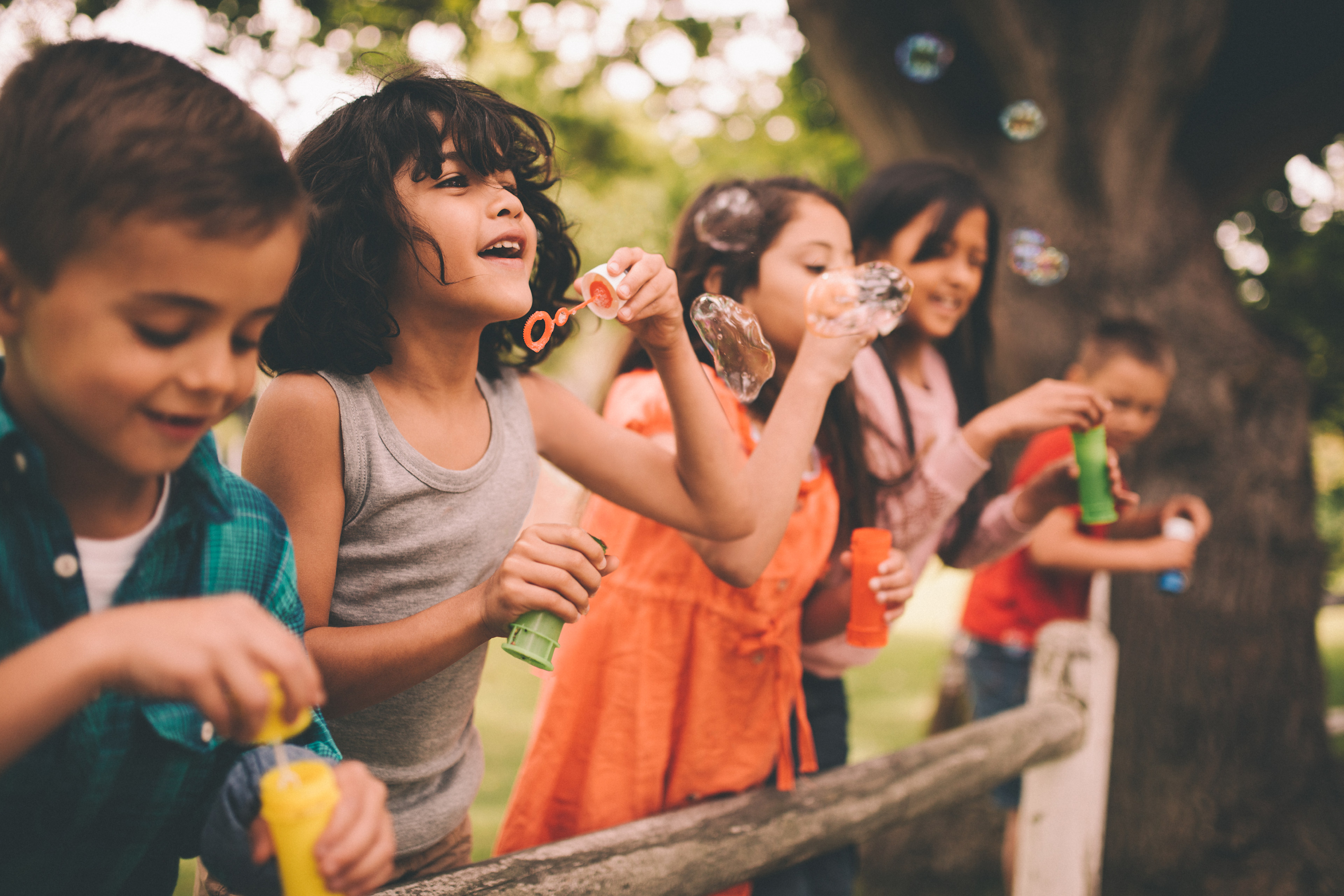 A group of young children playing with bubbles in a lush, outdoor setting with trees and greenery in the background.