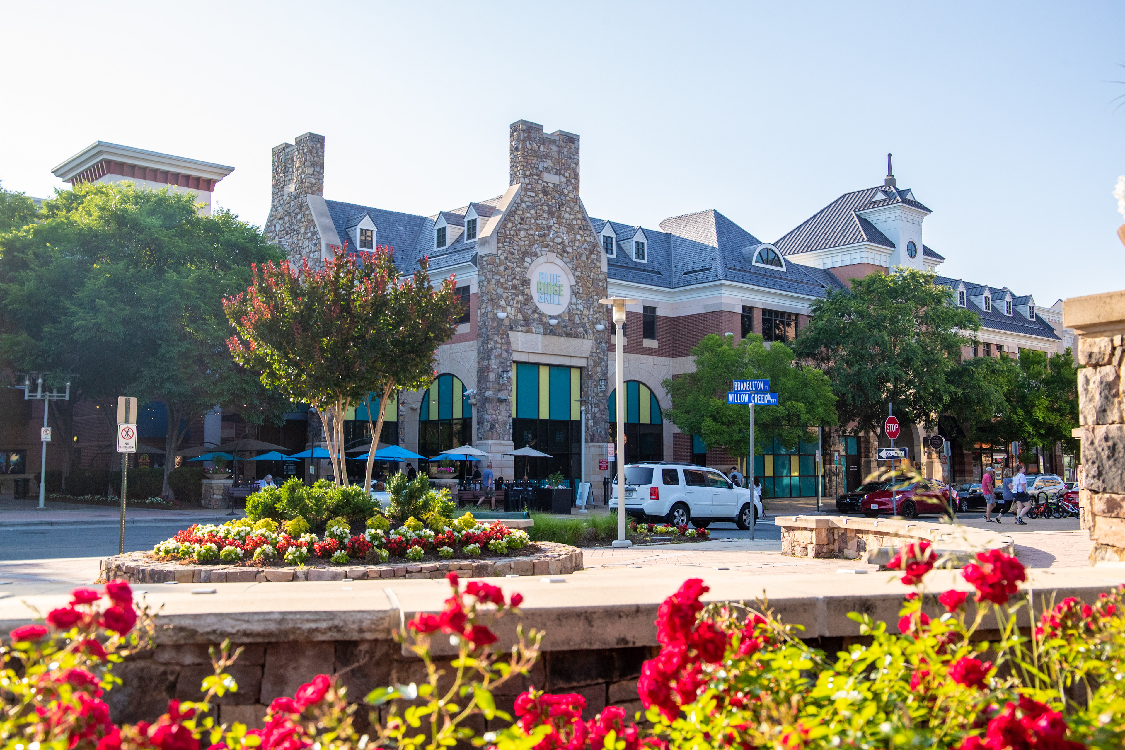 A vibrant and colorful urban scene with a stone fountain surrounded by blooming flowers in the foreground, and a collection of distinctive stone and brick buildings in the background.