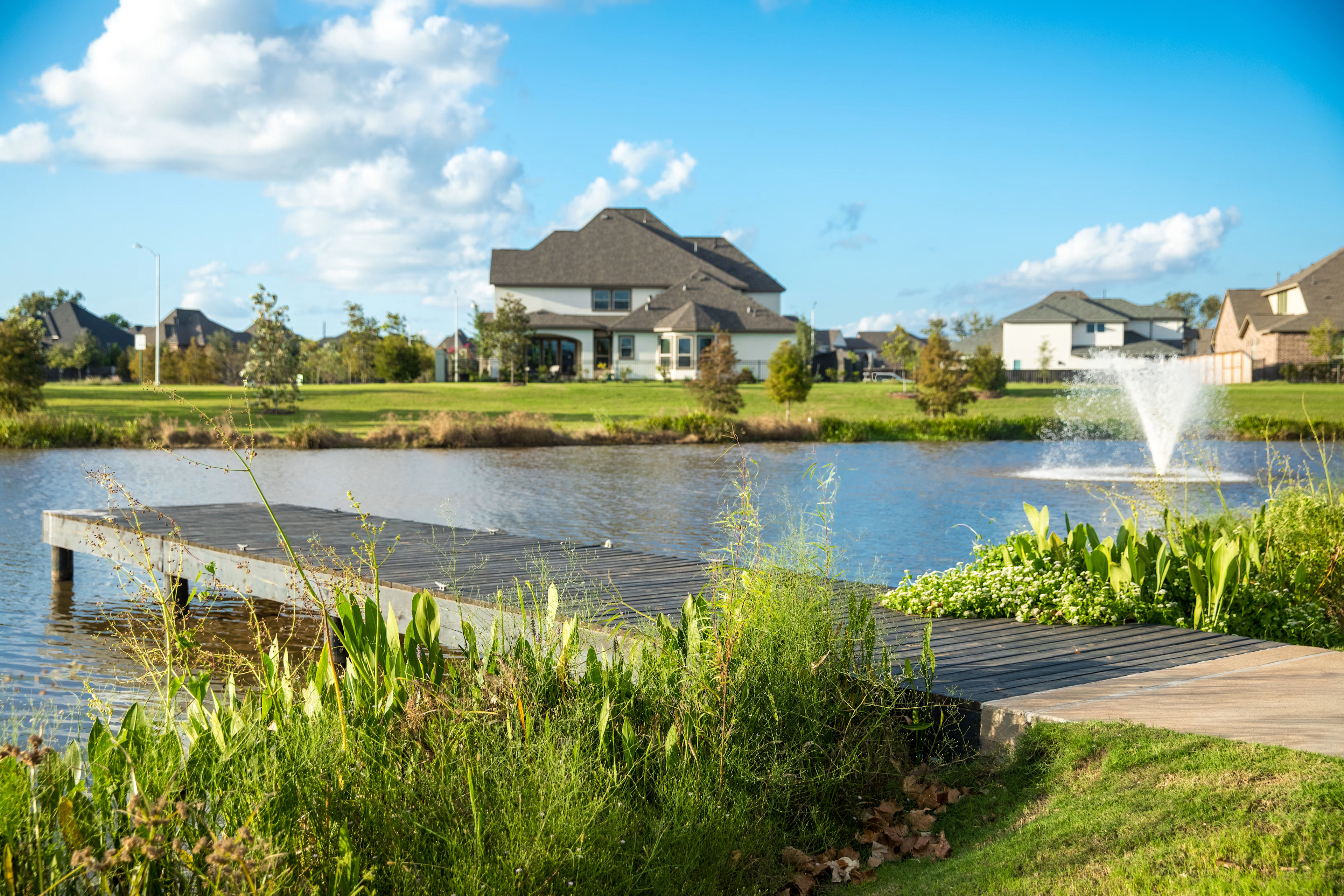A picturesque suburban neighborhood with a large house overlooking a tranquil pond, surrounded by lush greenery and a wooden dock in the foreground.