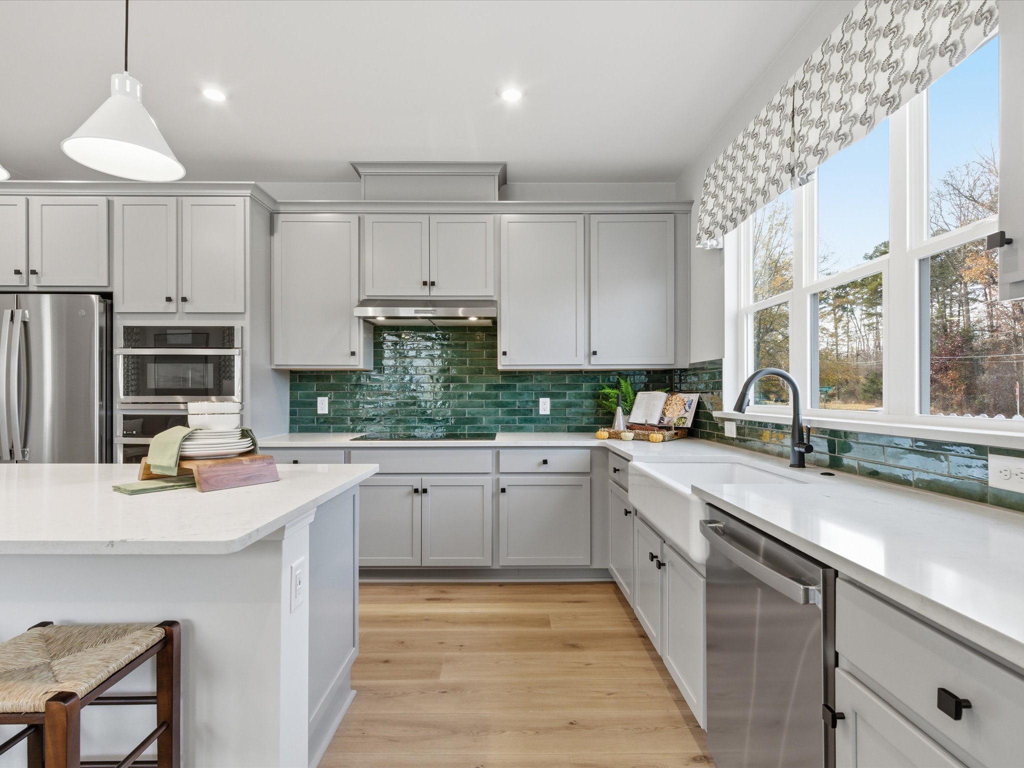 A modern, well-lit kitchen with gray cabinets, white countertops, and a green tile backsplash, featuring a large window overlooking a scenic outdoor view.