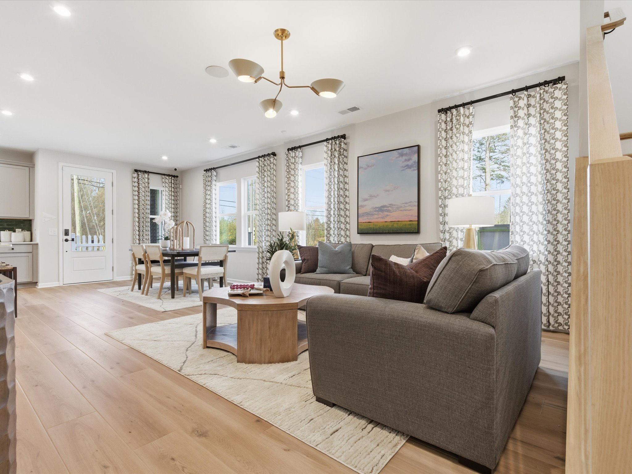 A spacious and well-furnished living room with a gray sofa, wooden coffee table, and a modern chandelier overhead, surrounded by large windows that allow natural light to flood the space.