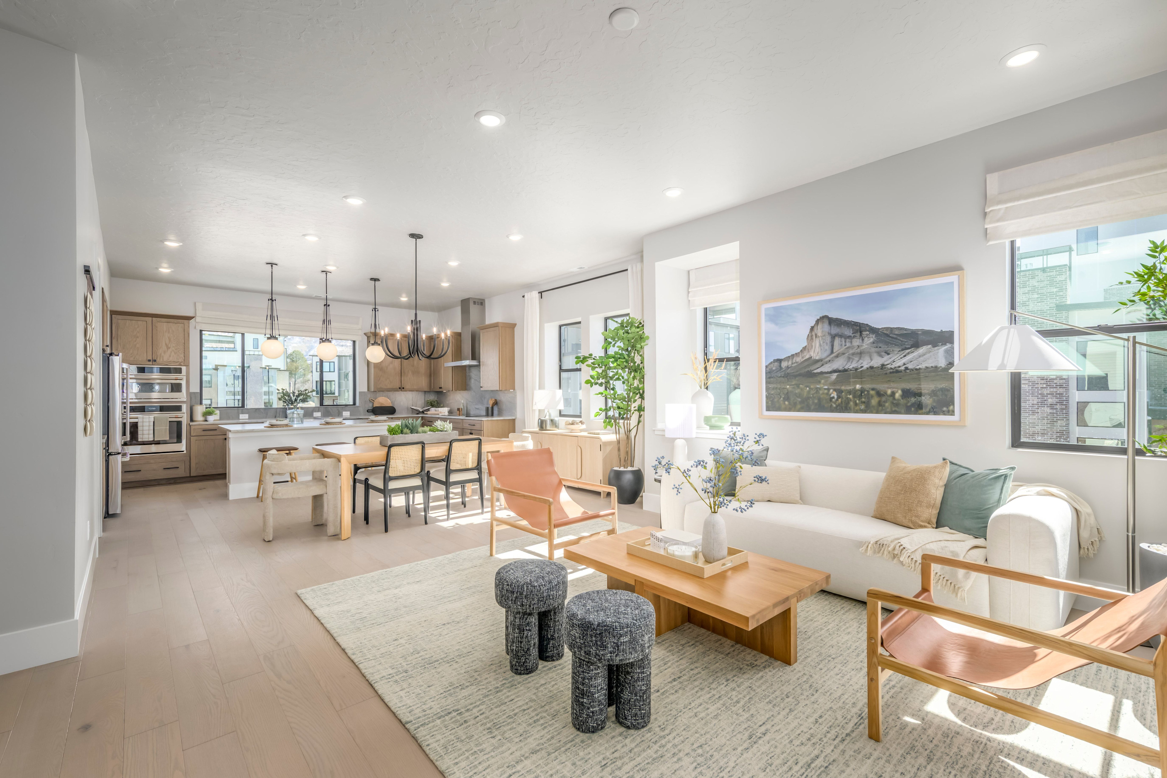 A modern and spacious open-concept living area with a kitchen, dining, and living room, featuring light-colored furniture, large windows, and a scenic landscape artwork on the wall.