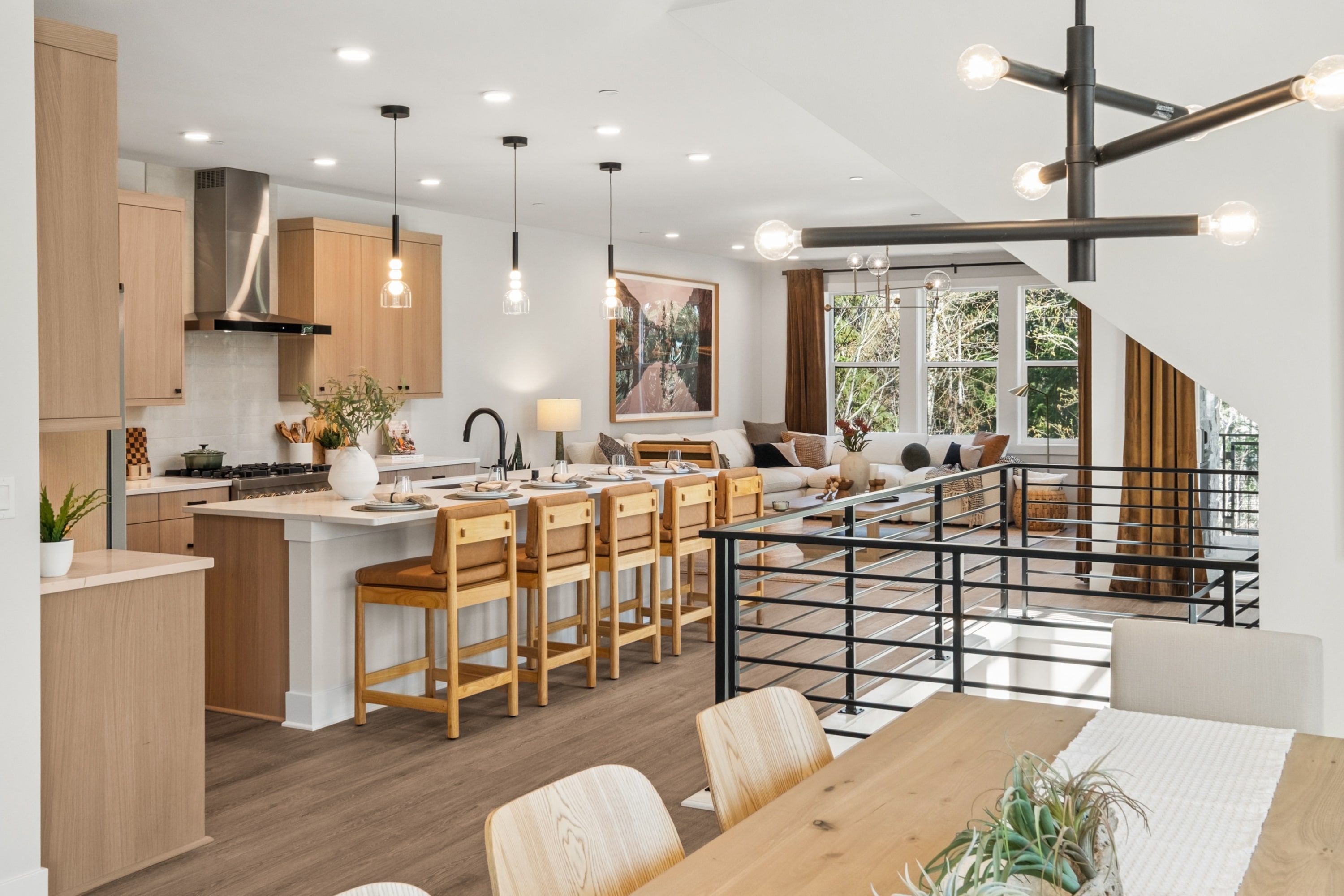 A modern and open-concept kitchen with a wooden dining table, bar stools, and large windows overlooking a lush outdoor area.