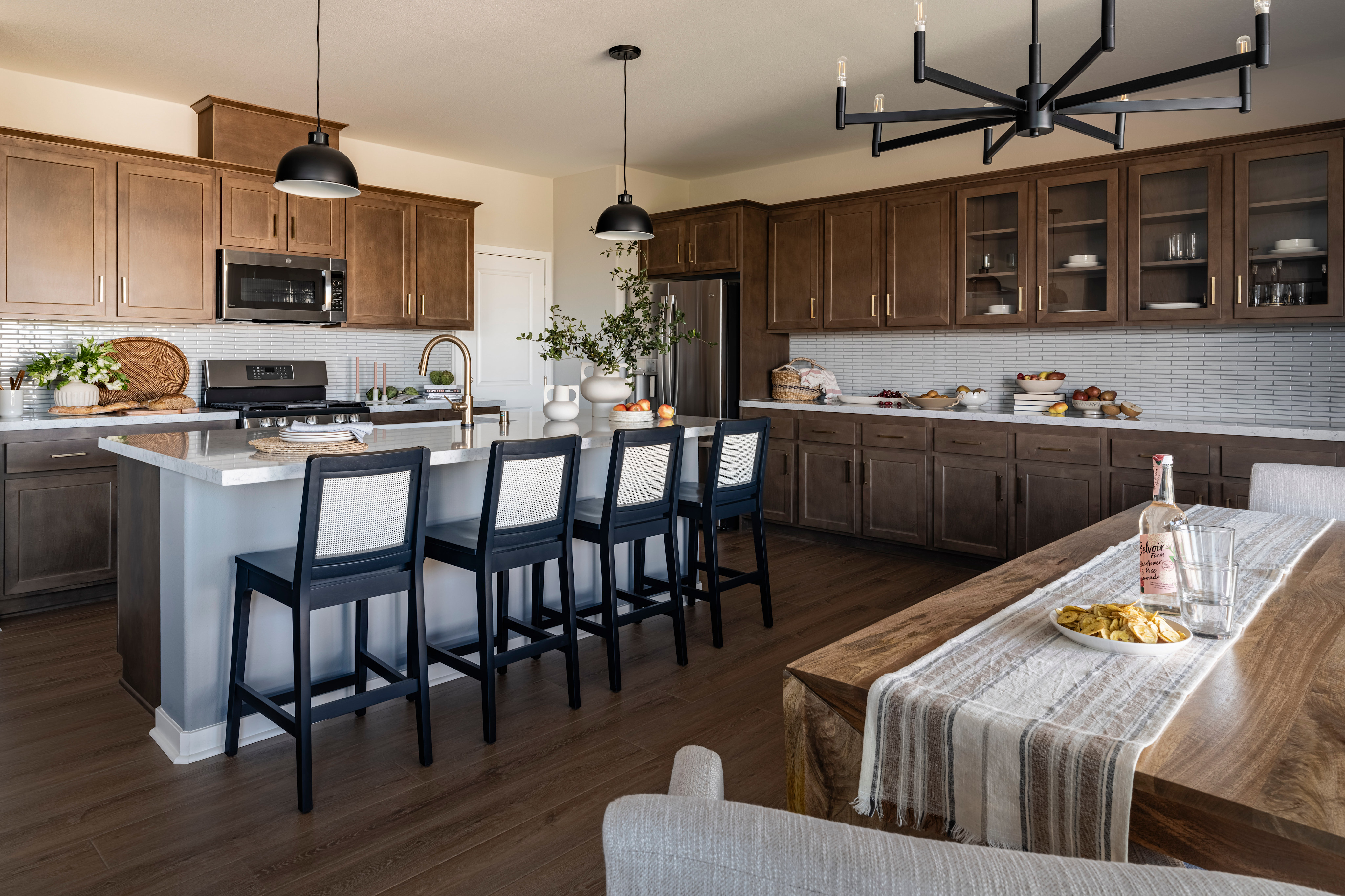 A modern and spacious kitchen with wooden cabinets, a large island with bar stools, and a chandelier overhead, set against a backdrop of a dining area with a table and chairs.