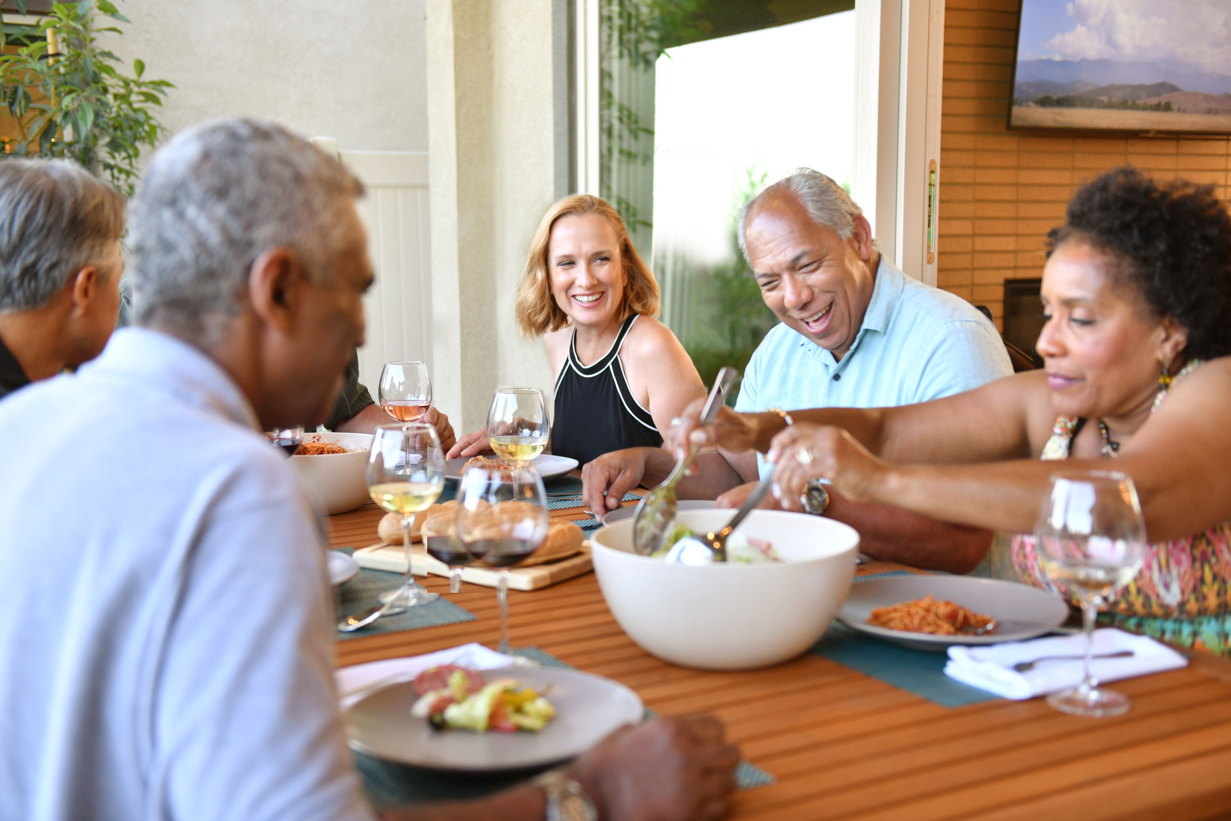 A group of people enjoying a meal together outdoors on a wooden deck, surrounded by lush greenery and a scenic background.
