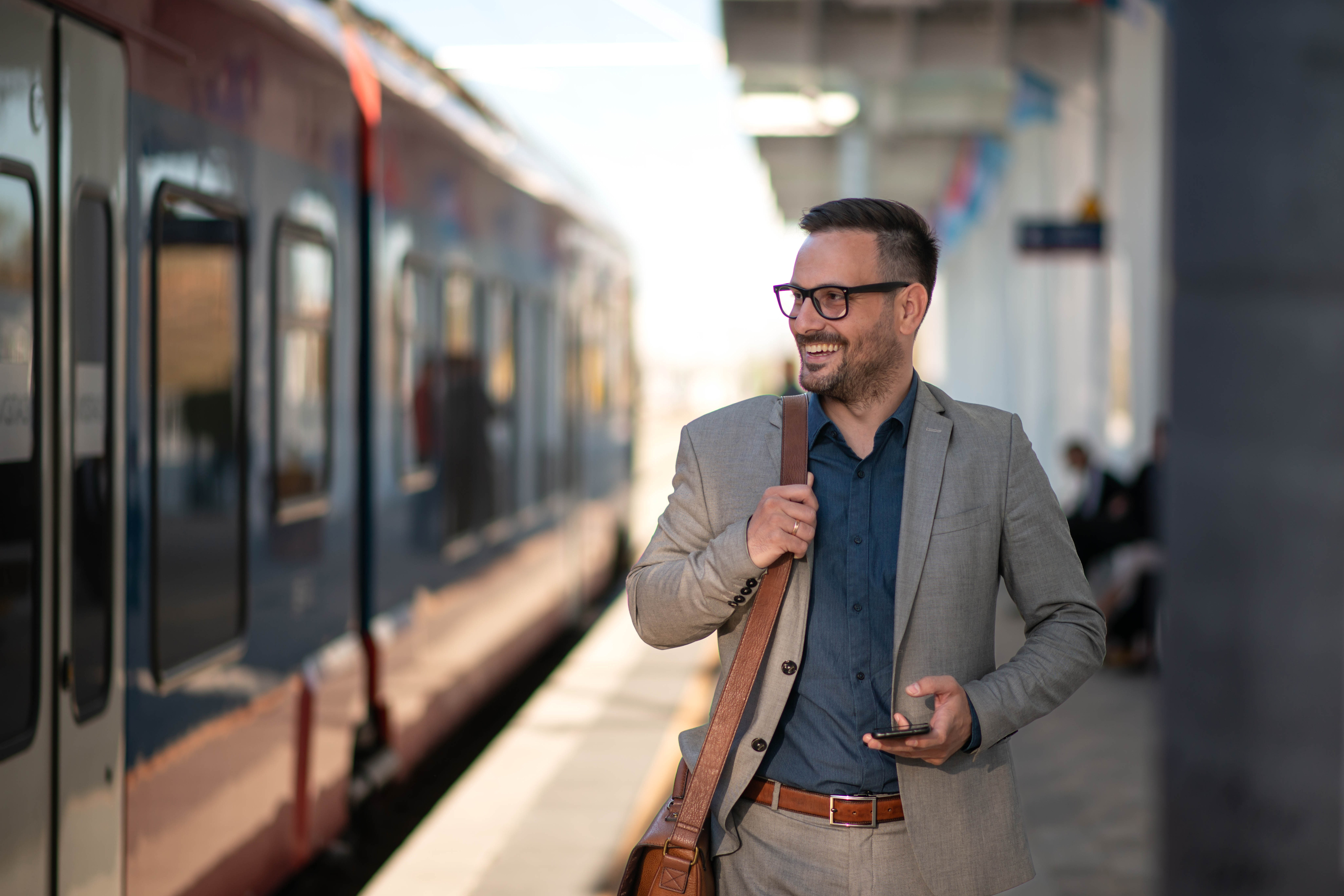A man in a gray suit and glasses stands on a train platform, holding a smartphone and smiling.