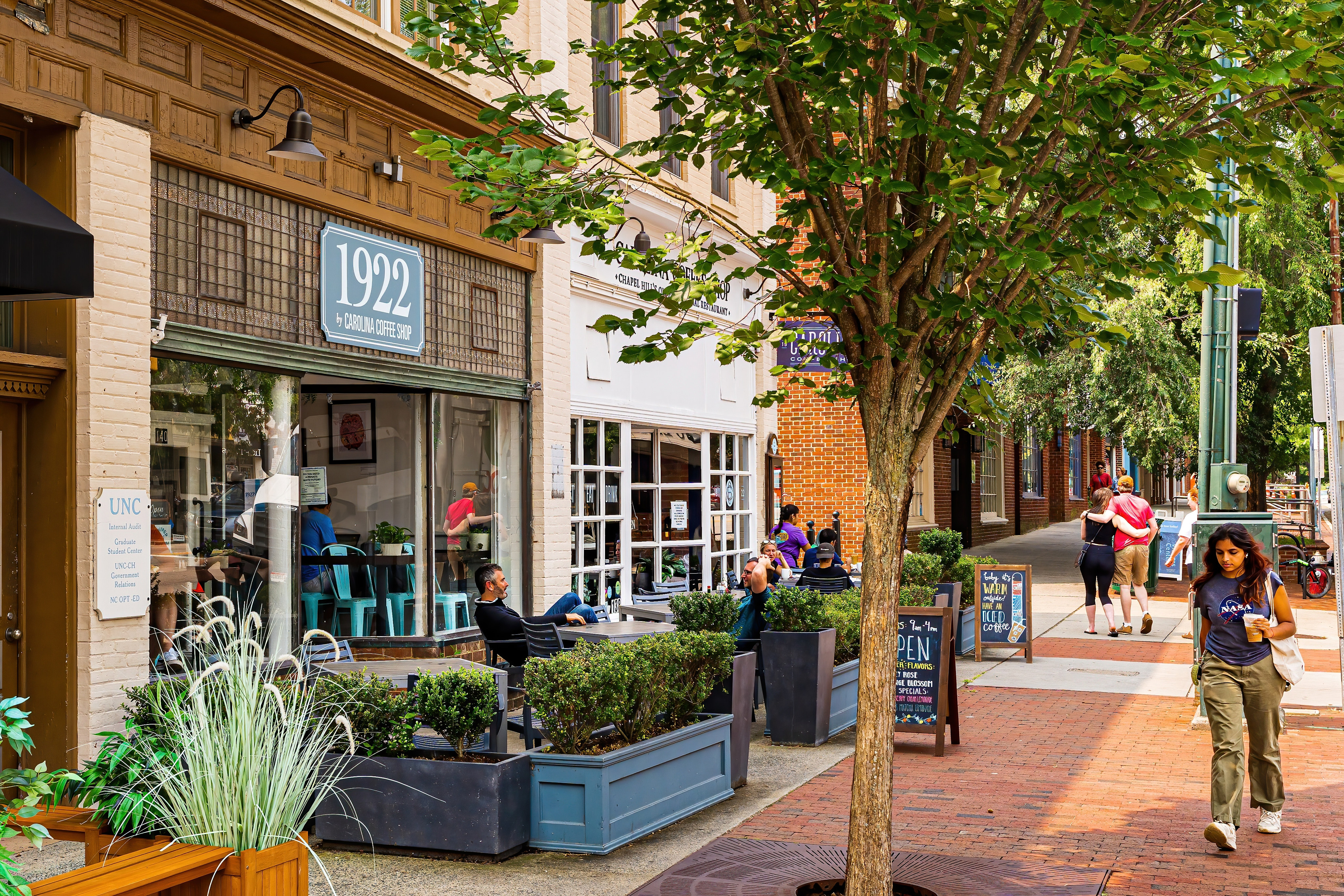 A bustling urban street scene with a variety of shops, pedestrians, and a large tree providing shade along the sidewalk.