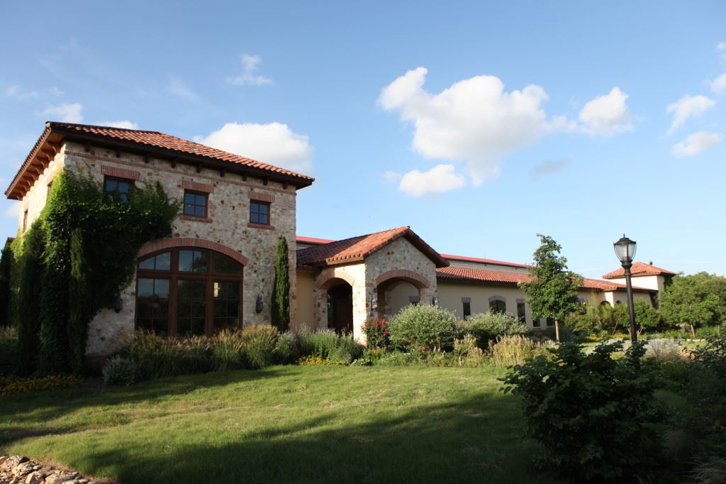 A stately Mediterranean-style villa with a red tile roof sits amidst a lush, well-manicured garden, under a bright, cloudless sky.
