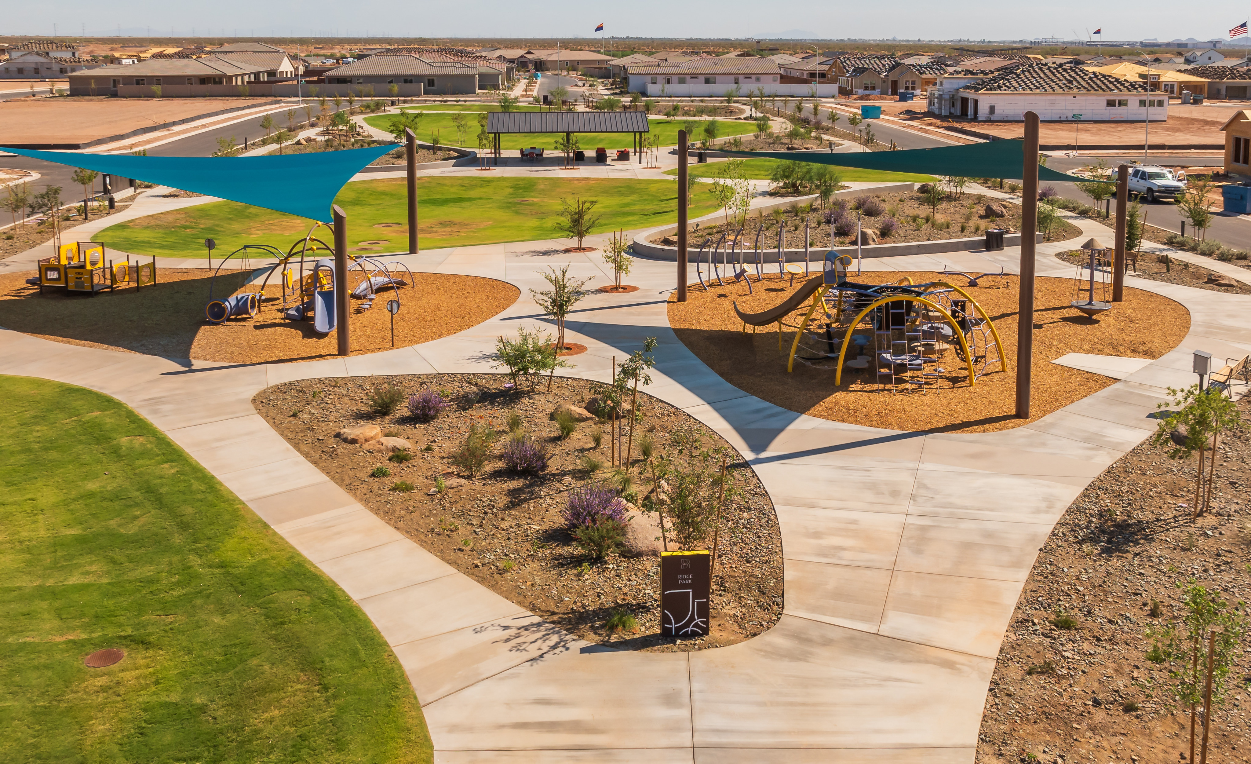 A vibrant and well-designed outdoor recreational area with various play structures, shaded seating areas, and lush landscaping, set against a backdrop of residential buildings.