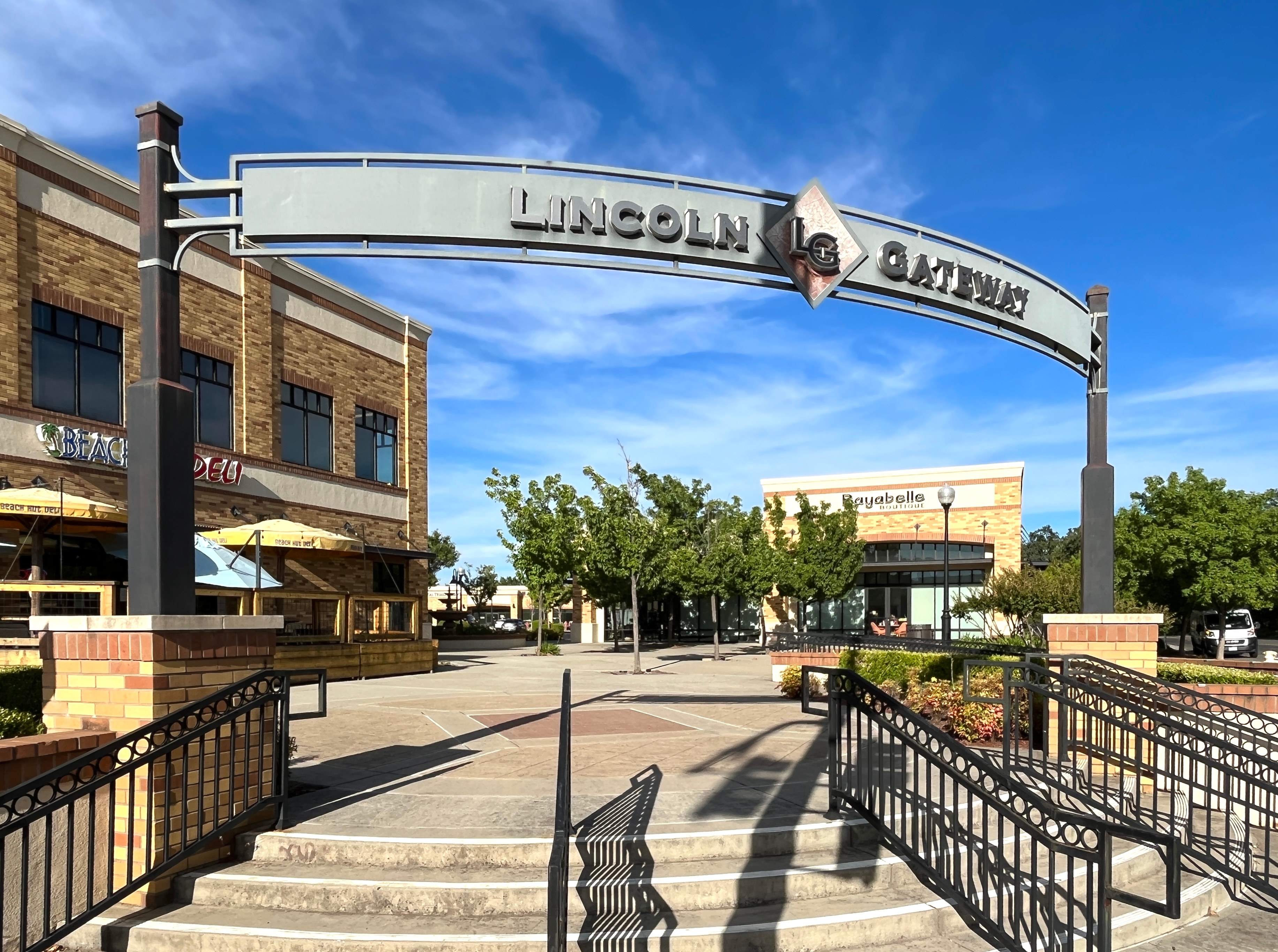 A brick-lined walkway leads to a large archway sign that reads "Lincoln Center" against a backdrop of blue sky and surrounding buildings.