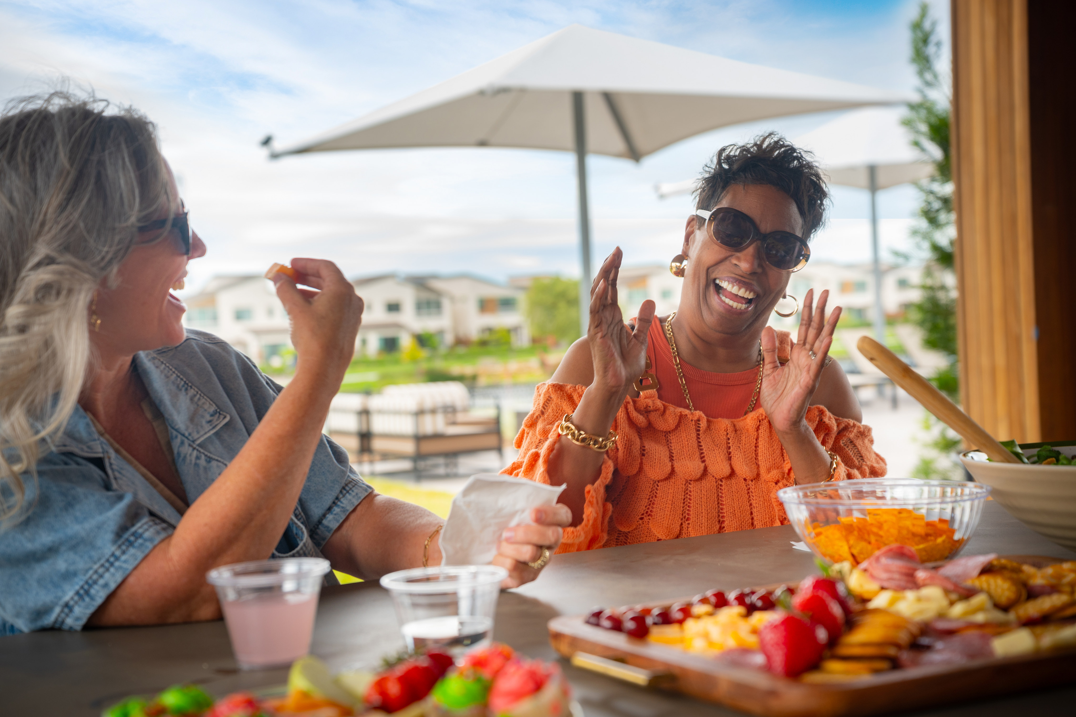 Two women laugh heartily at a table laden with snacks, with a modern outdoor patio and distant houses under a bright sky in the background.