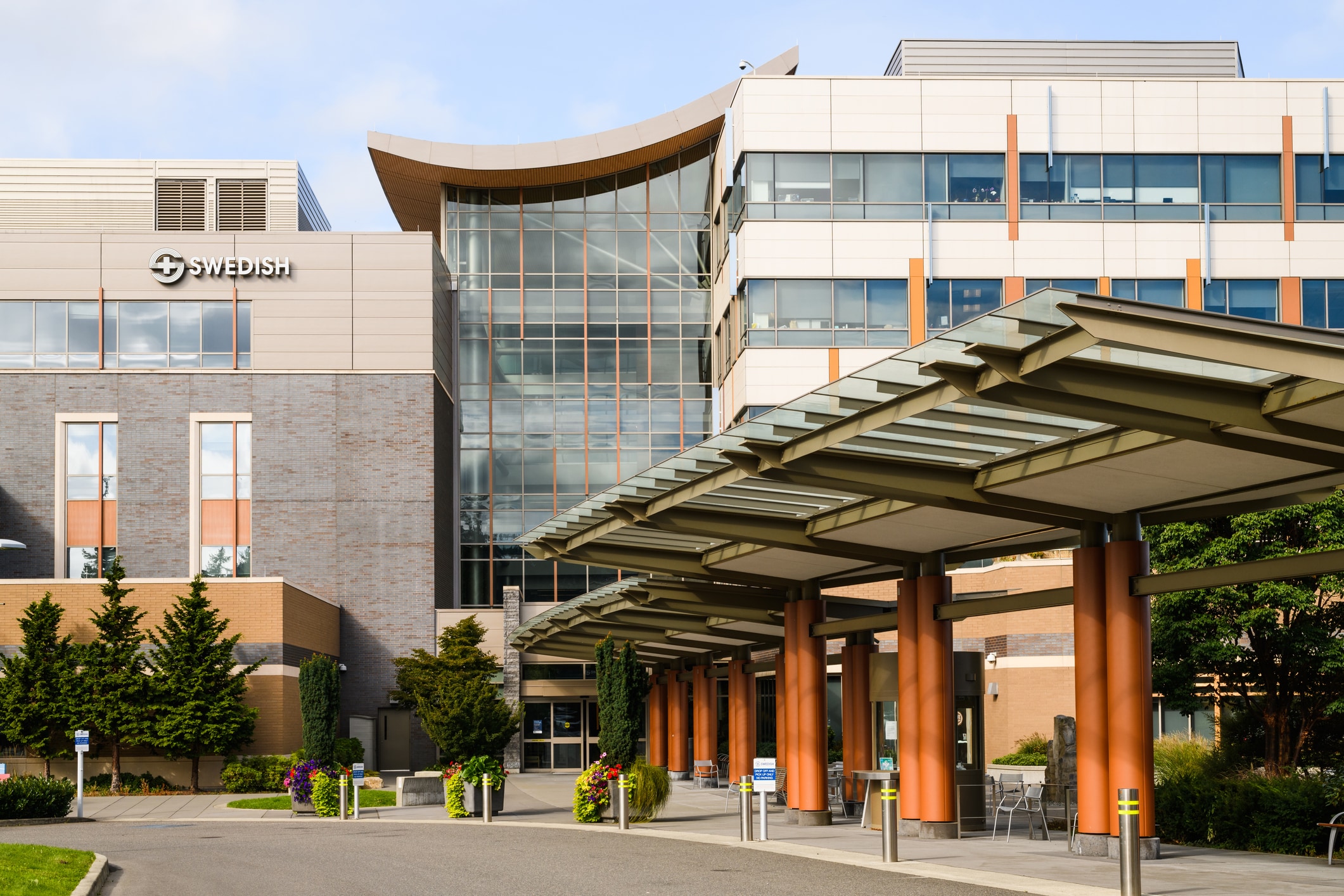 A modern hospital building with a covered walkway in the foreground and trees and landscaping surrounding it.