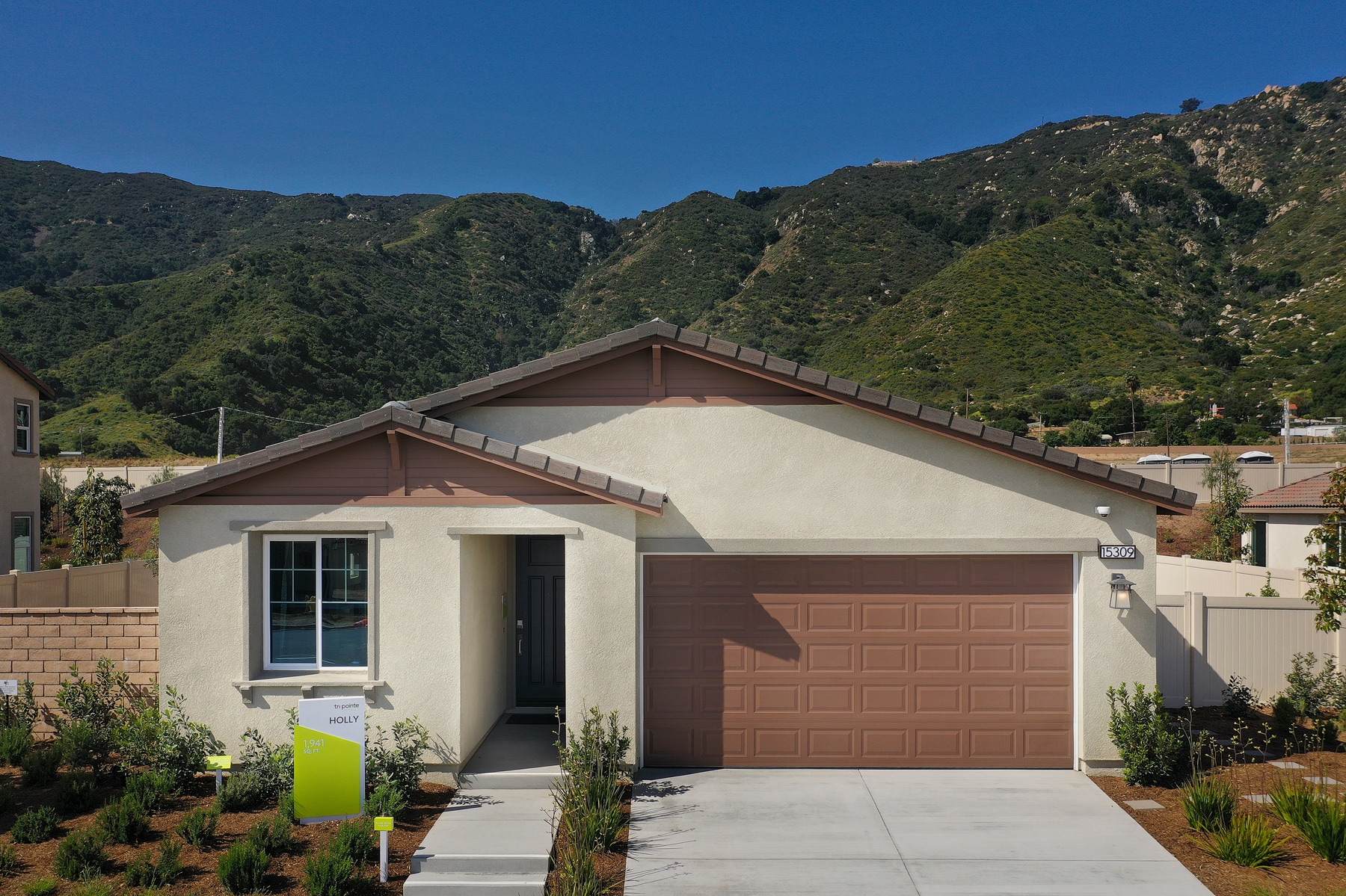 A single-story residential house with a brown garage door and a small yard in the foreground, set against a backdrop of lush, green mountains.