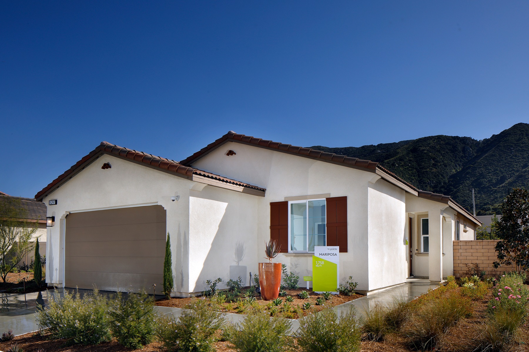 A single-story white house with red-tiled roofs sits in a lush, grassy yard surrounded by mountains in the background under a clear blue sky.