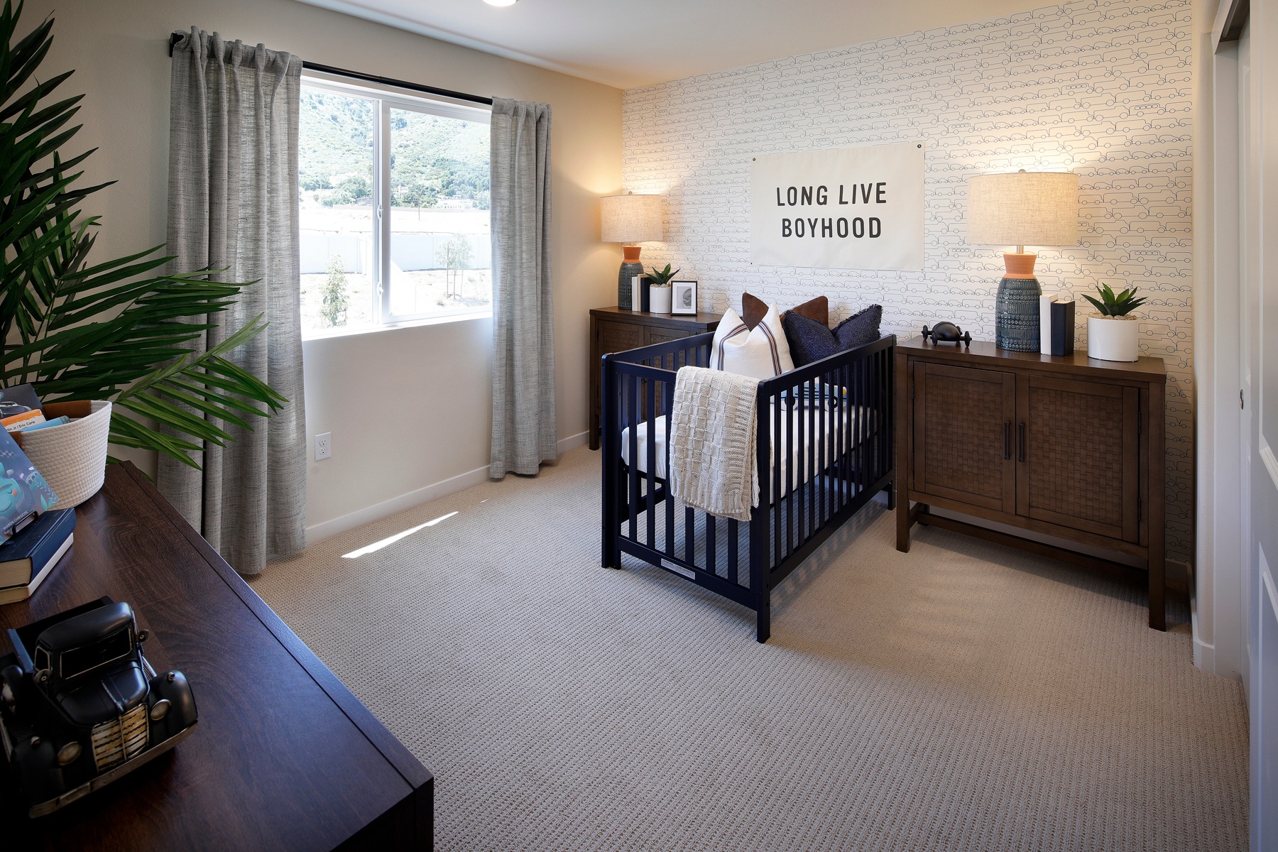 A cozy nursery with a black crib, a wooden dresser, and a wall sign that reads "Long Live Boyhood", surrounded by lush greenery and natural light.