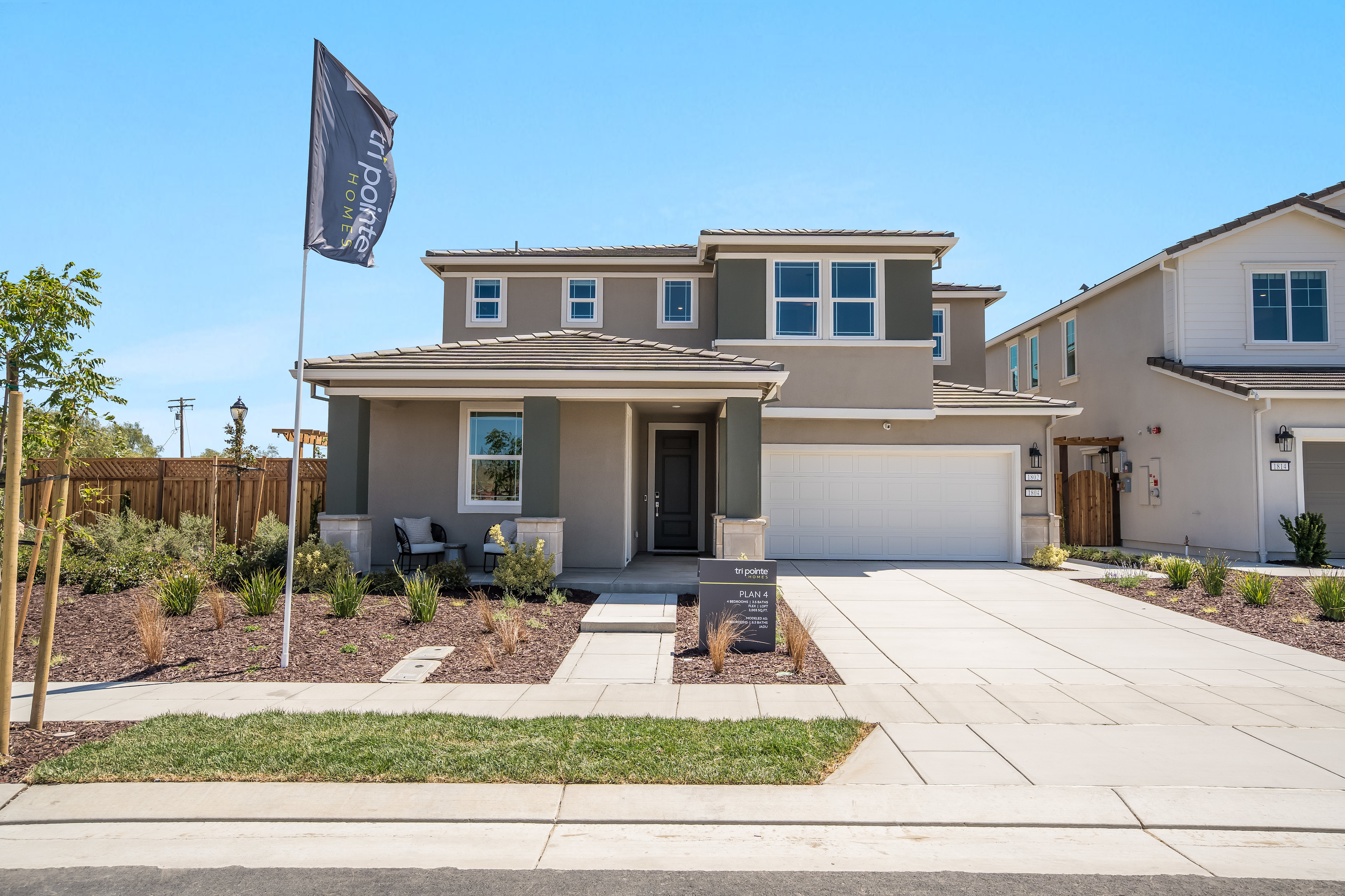 A two-story residential house with a garage, surrounded by a landscaped yard and a paved driveway, set against a clear blue sky.