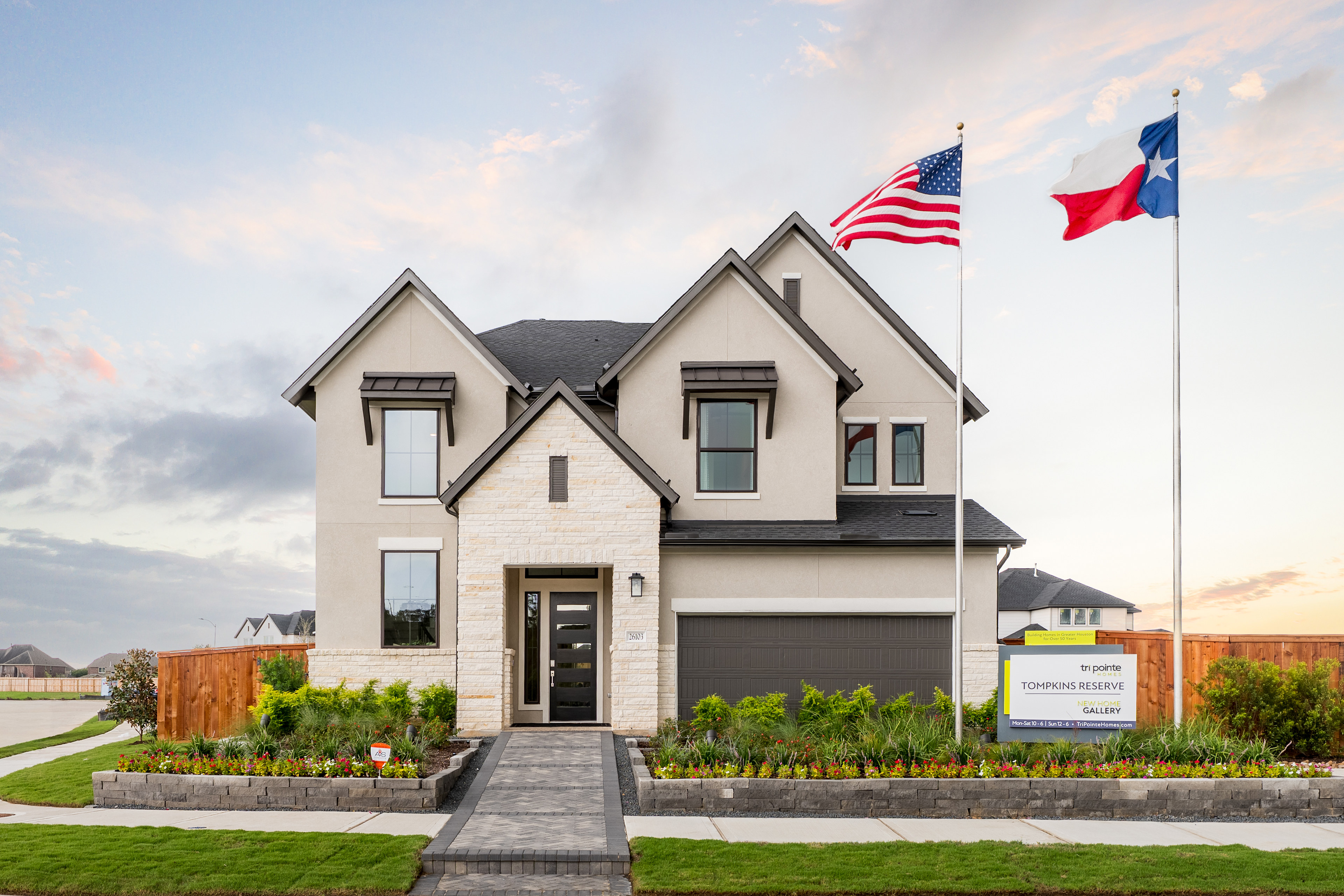 A two-story residential house with a garage, surrounded by a well-manicured lawn and landscaping, with an American flag and a Texas state flag flying in the foreground.