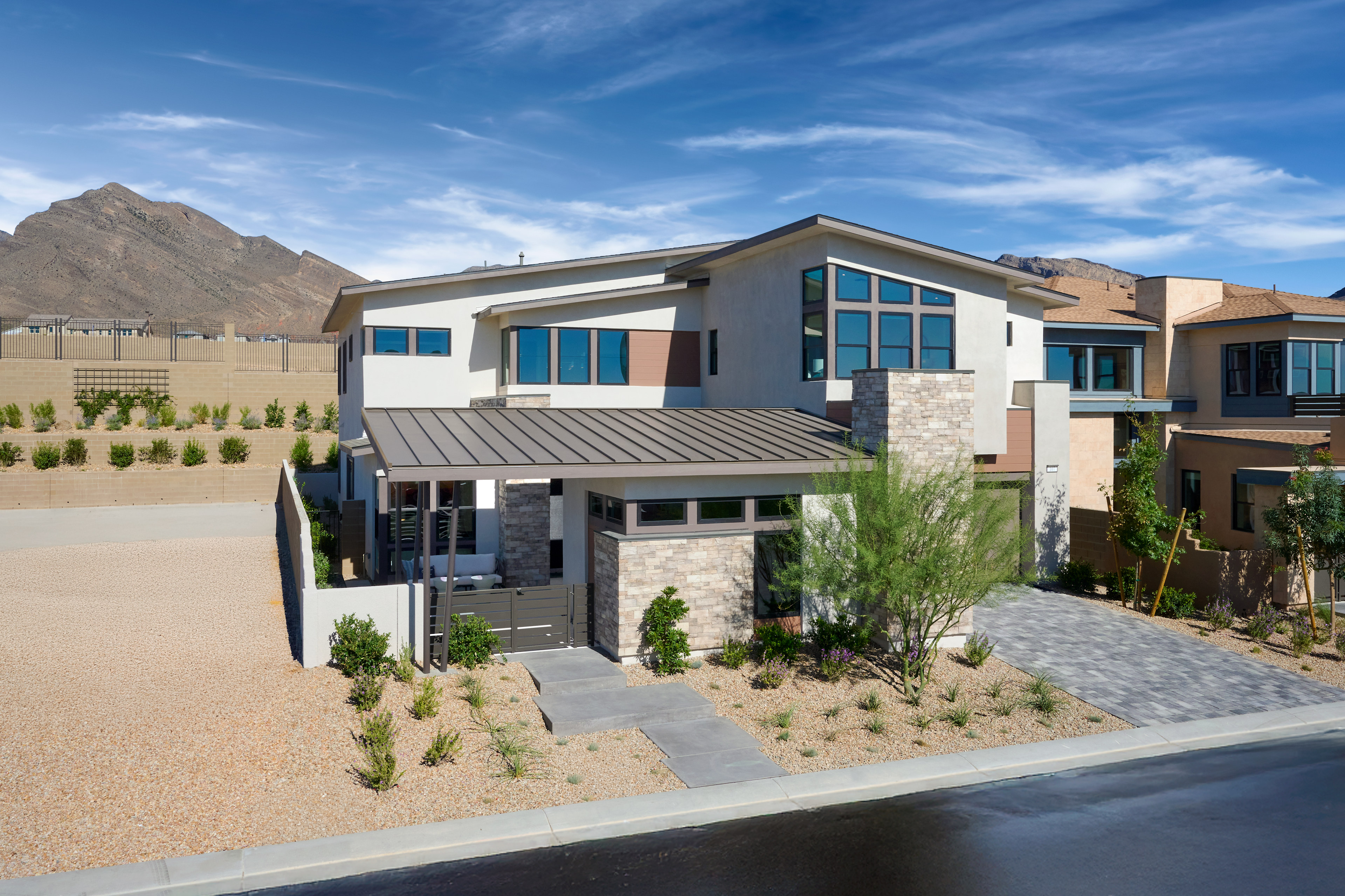A modern, two-story house with a stone exterior, large windows, and a metal roof sits in the foreground, surrounded by a desert landscape with mountains in the background.