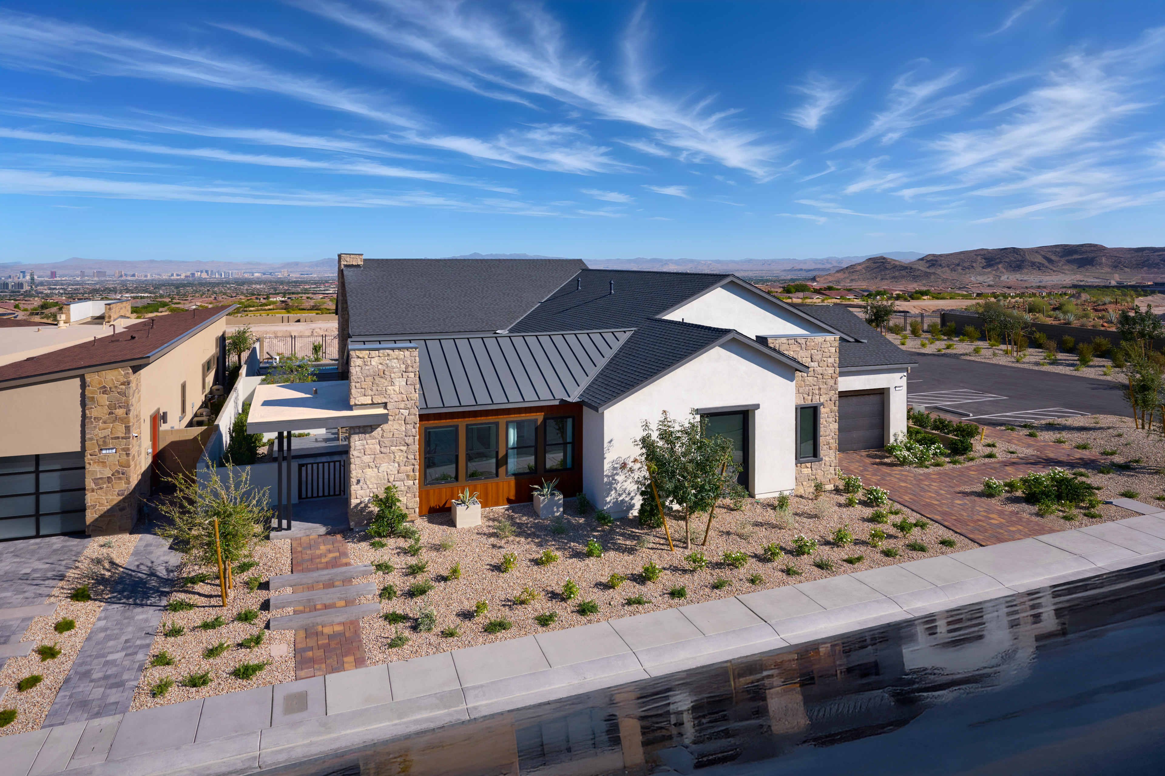 A modern, single-story house with a gray roof and white exterior sits in a desert landscape, surrounded by a paved driveway and lush landscaping, with a clear blue sky and wispy clouds overhead.