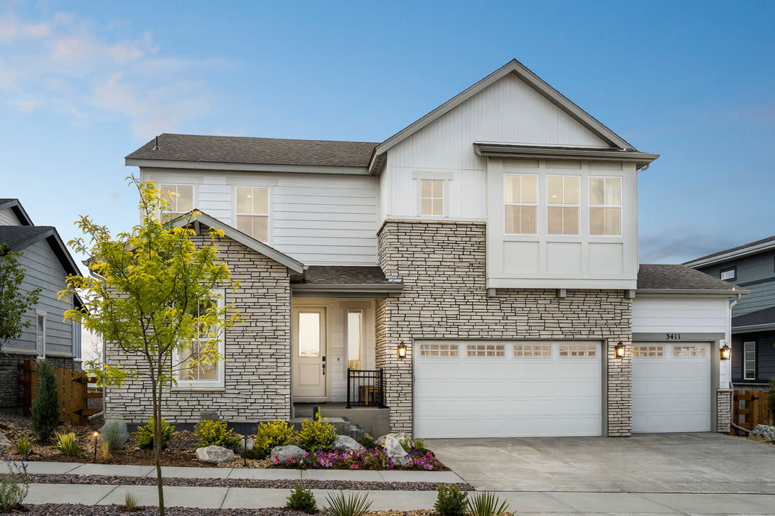 A two-story residential house with a white exterior, stone accents, and a garage door in the foreground, set against a backdrop of a clear blue sky and lush greenery.