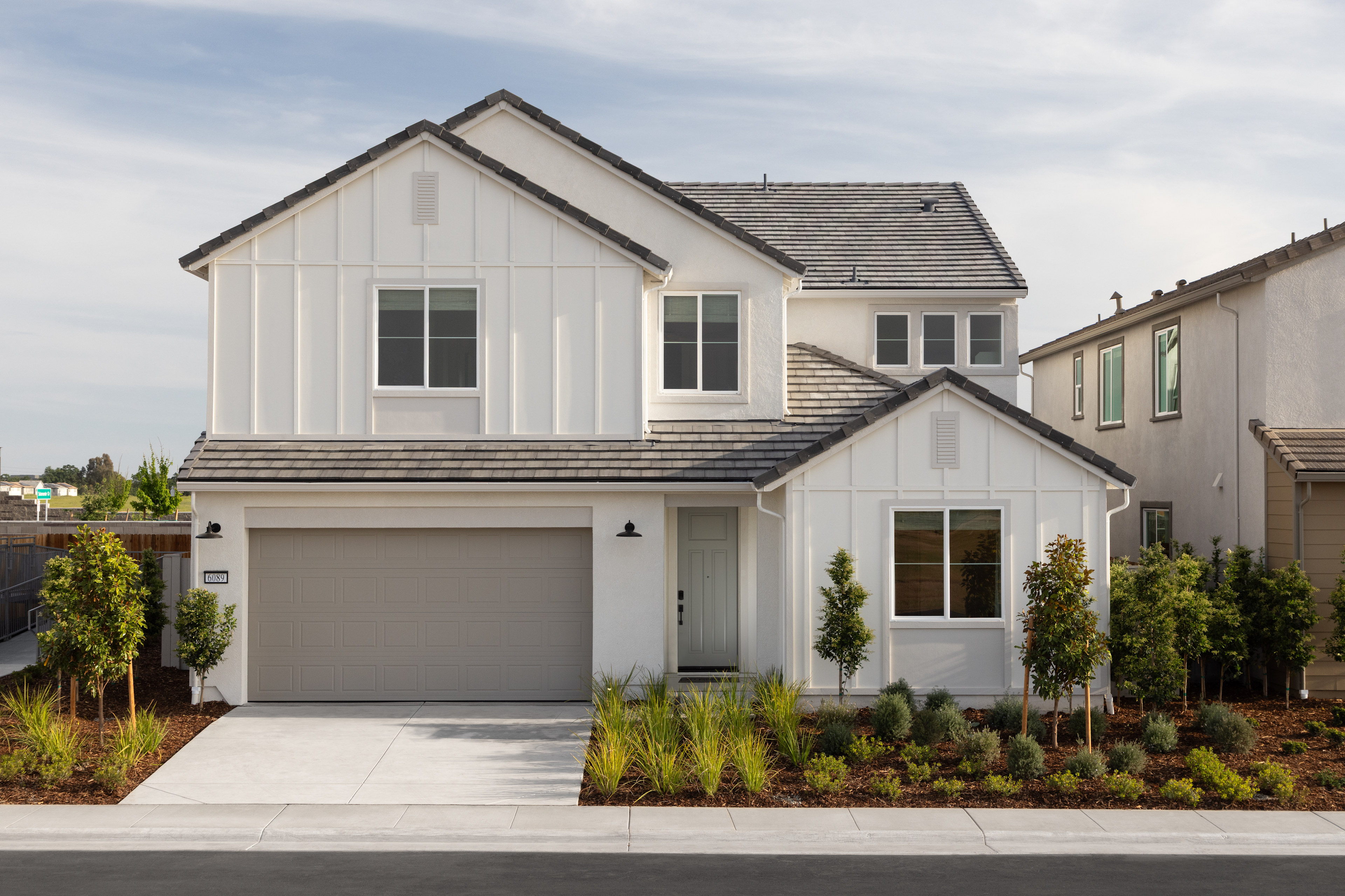 A two-story white house with a gray roof, surrounded by landscaped greenery, stands in the foreground against a cloudy sky in the background.