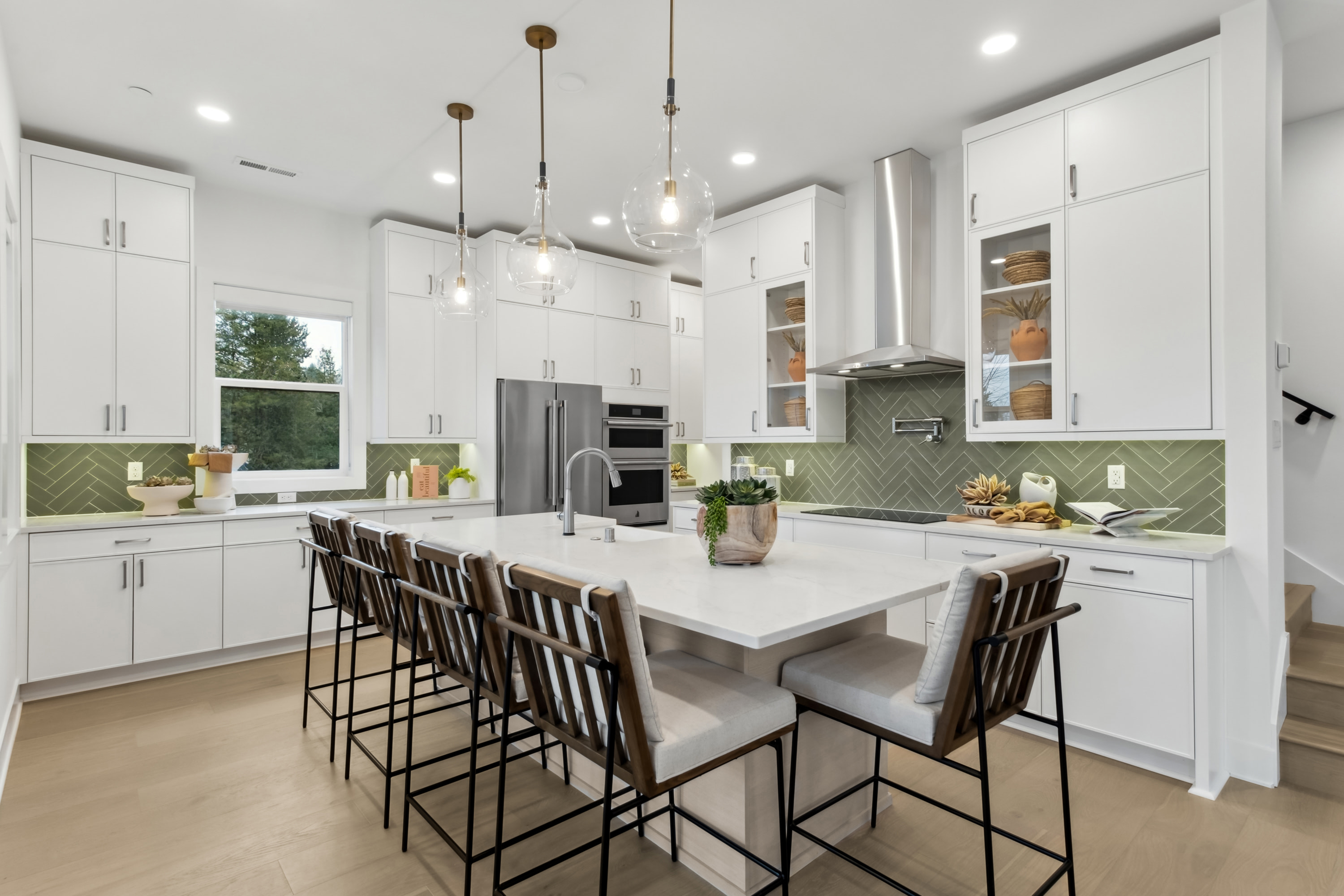 A modern, bright kitchen with white cabinets, a large island with bar stools, and a green tile backsplash, complemented by pendant lighting and a view of the outdoors through a window.