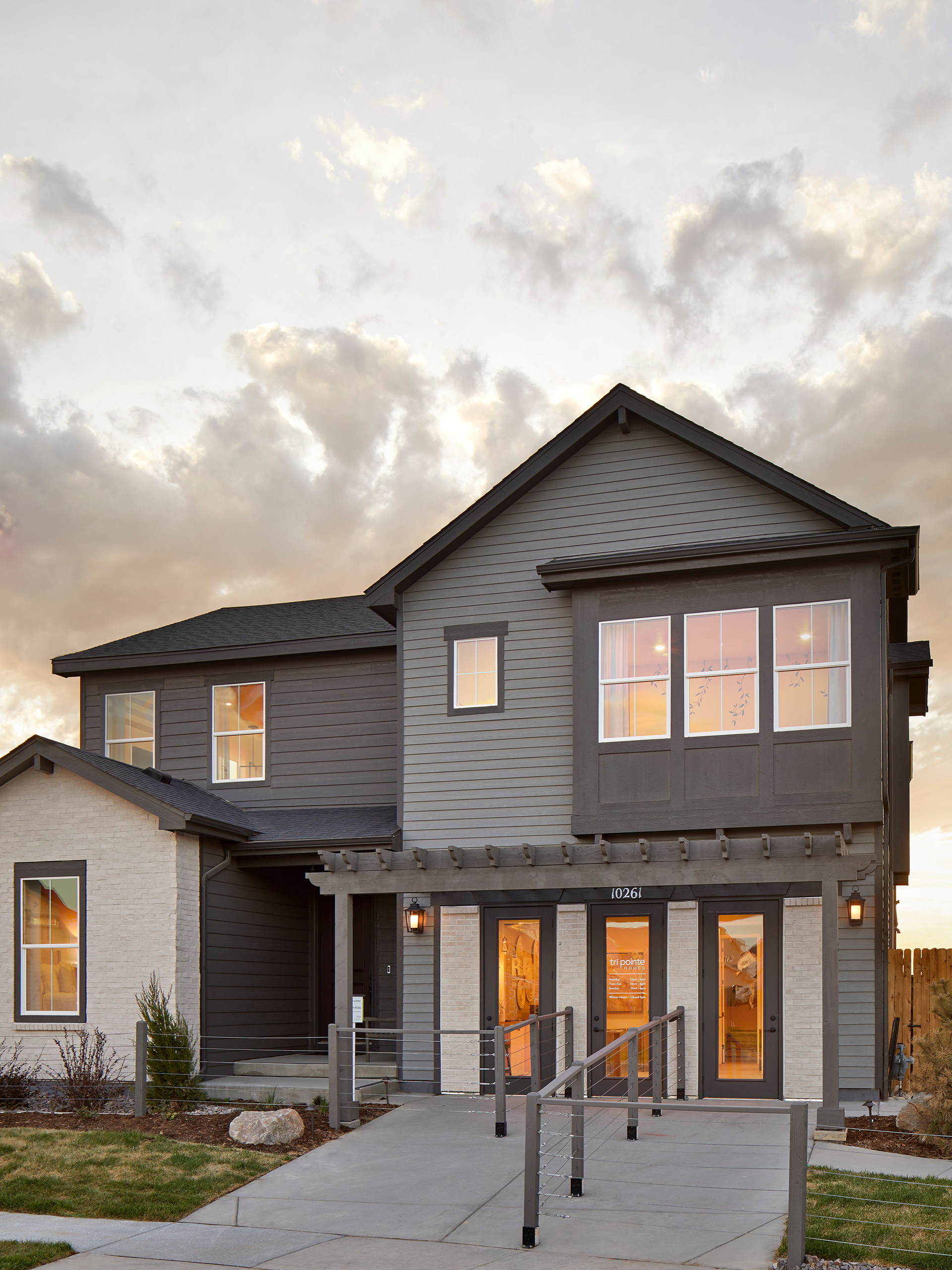 A two-story residential building with a porch and large windows, set against a cloudy sky with a grassy foreground.