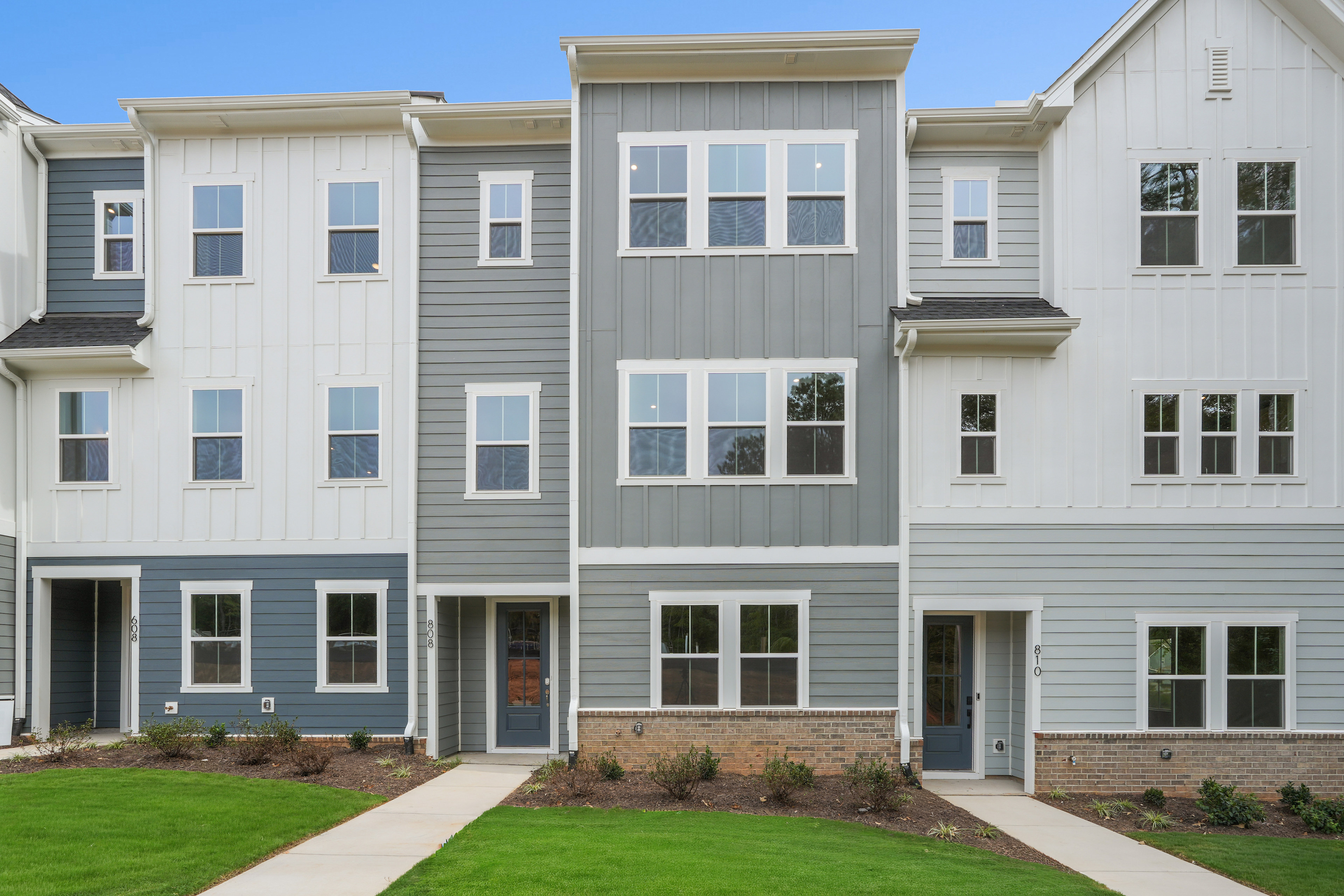 The image depicts a modern, multi-story townhouse complex with a well-manicured lawn in the foreground.