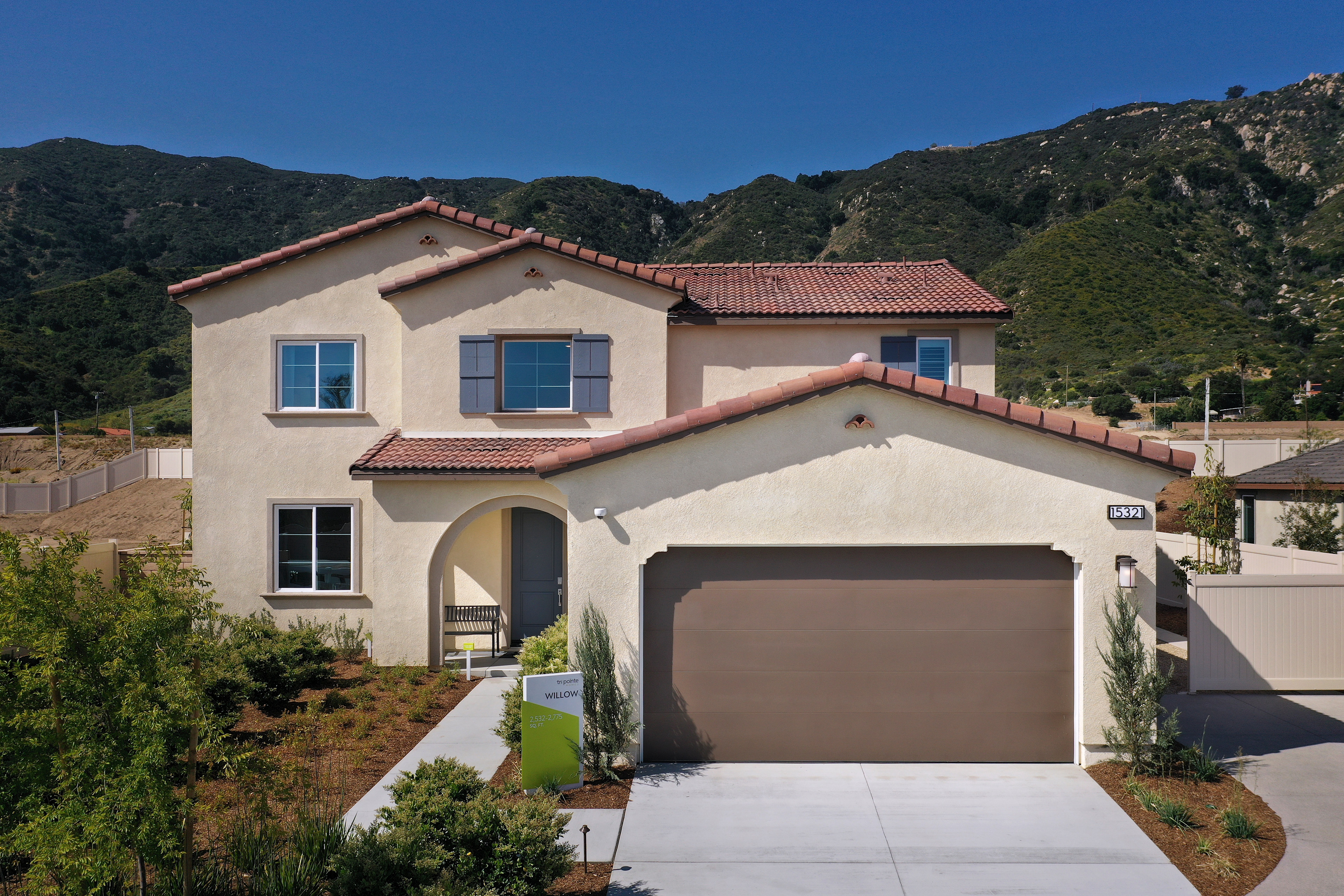 A two-story Mediterranean-style house with a red tile roof, surrounded by lush greenery and set against a backdrop of rugged mountains.