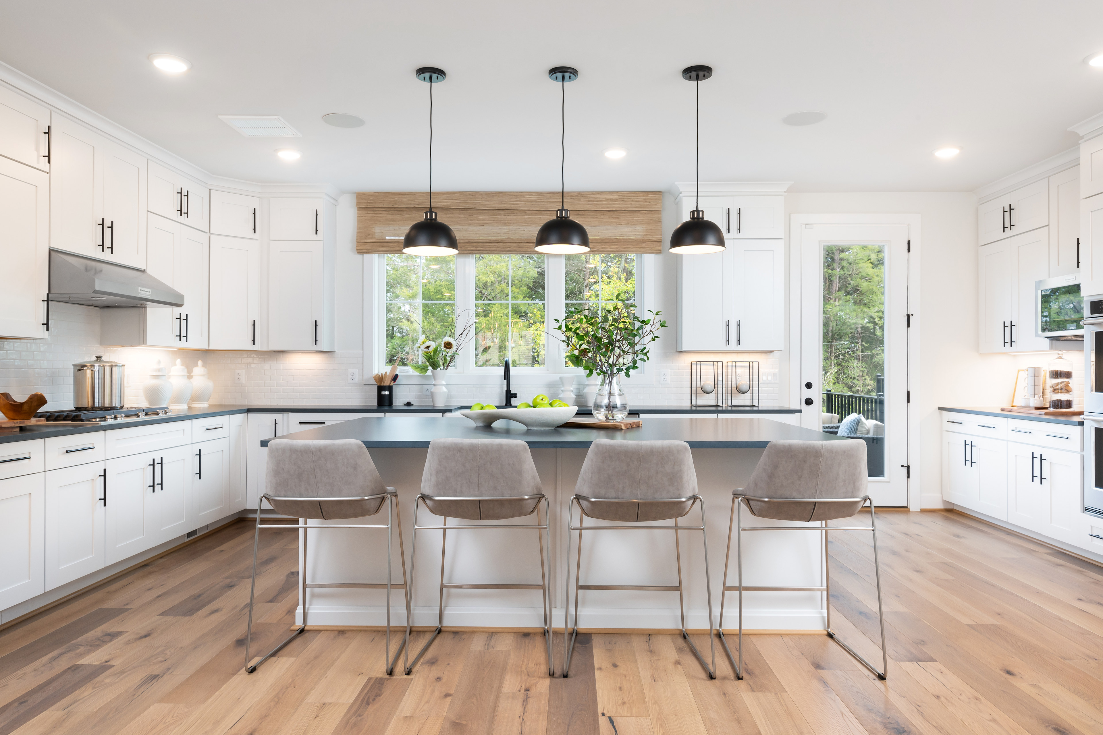 A modern, bright, and spacious kitchen with white cabinets, a large island with bar stools, and pendant lights hanging above, overlooking a scenic outdoor view through the window.